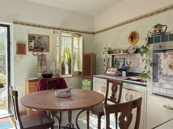 Pannello di cucina rustica con tavolo rotondo in legno, sedie, e dettagli vintage, ambiente luminoso con finestre e accessori decorativi, atmosfera accogliente e tradizionale.