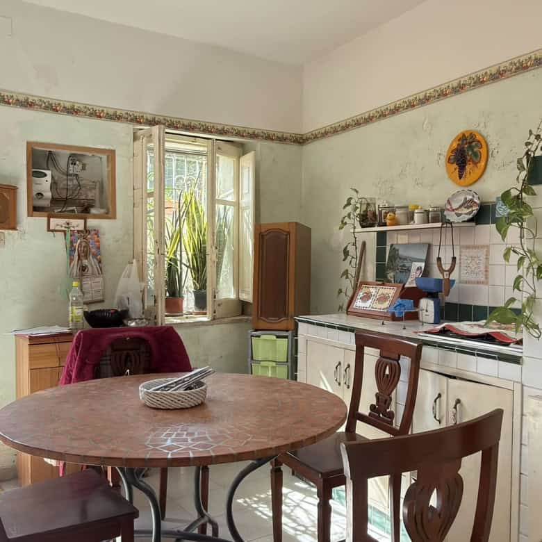 Pannello di cucina rustica con tavolo rotondo in legno, sedie, e dettagli vintage, ambiente luminoso con finestre e accessori decorativi, atmosfera accogliente e tradizionale.