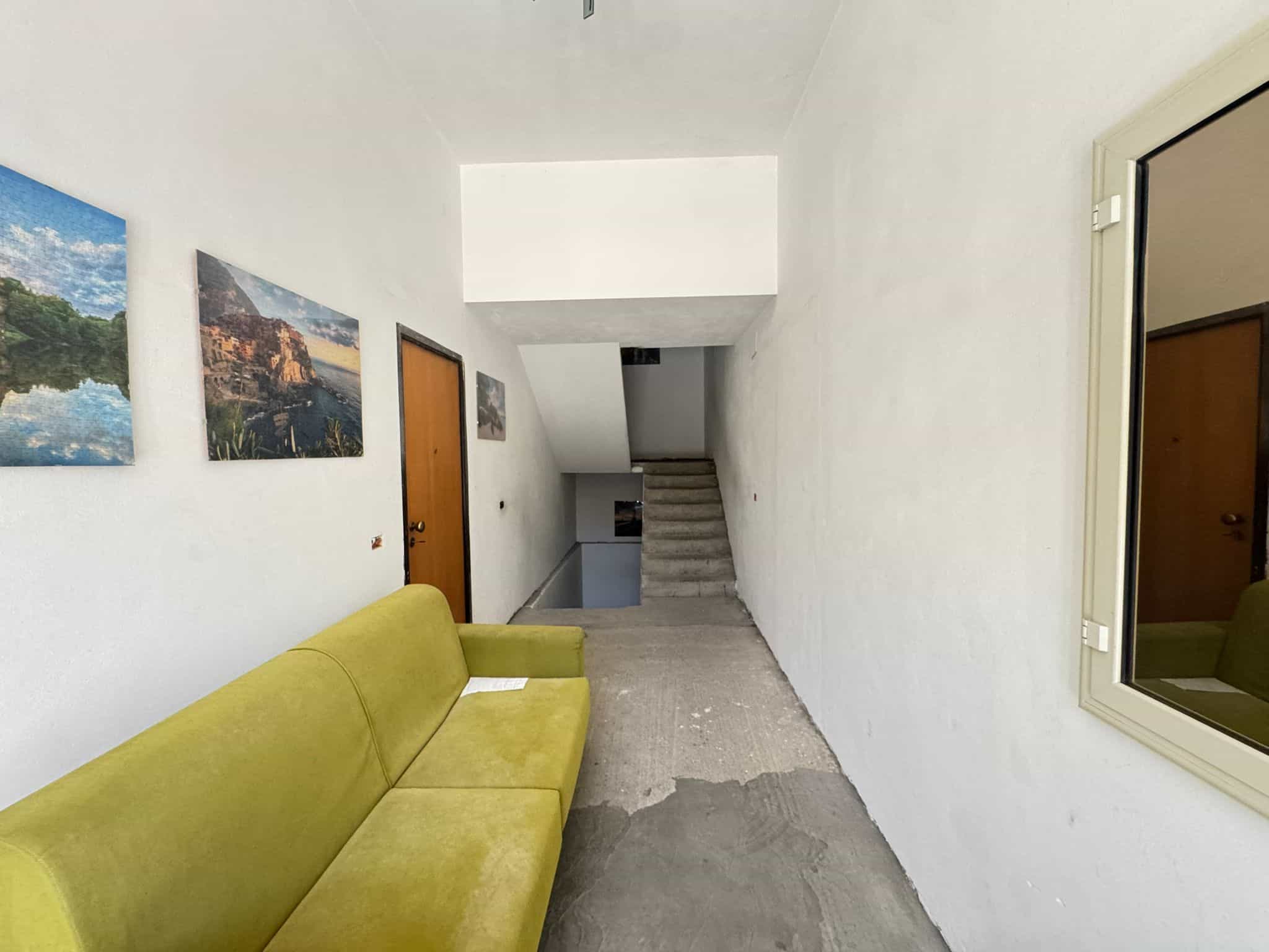 Sedia verde in un ingresso di casa con pareti bianche e quadri decorativi, vista verso le scale interne e una finestra riflessa nello specchio.