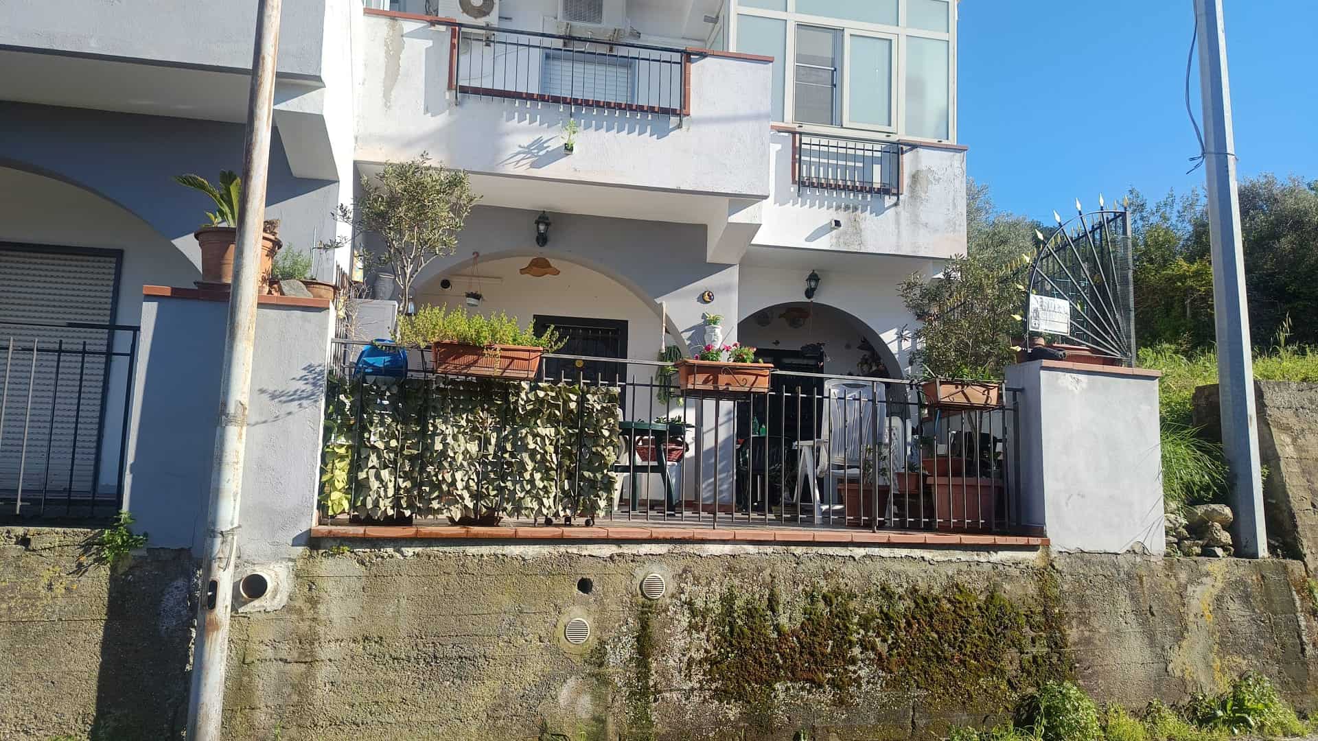 Balcone di una casa moderna con piante e arredi, vista su strada e cielo azzurro, in un quartiere residenziale.