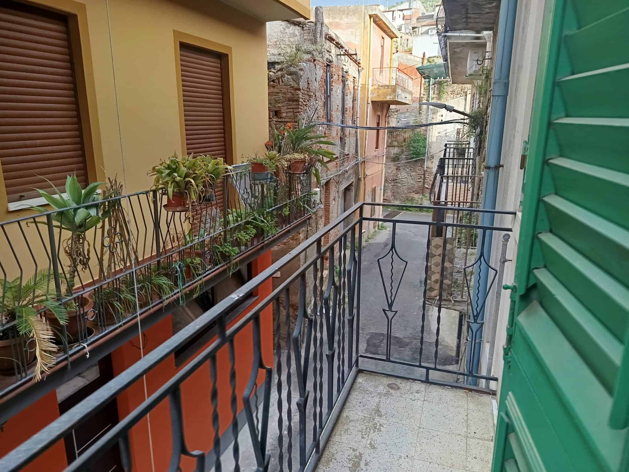 Balcone di un appartamento in un quartiere storico, con piante in vaso e ringhiera in ferro battuto, vista su stretta strada urbana con edifici colorati e dettagli architettonici tradizionali.