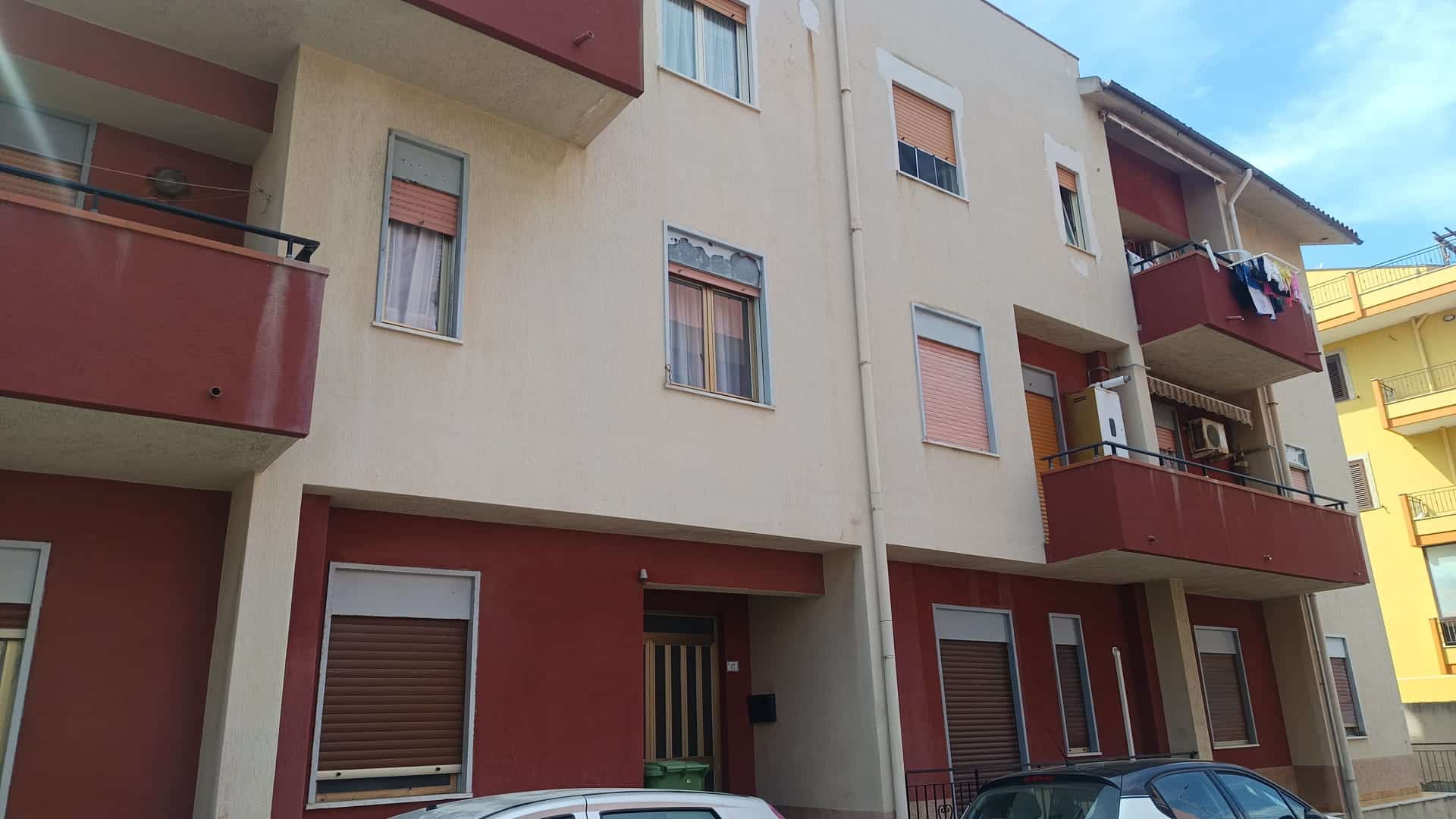 Ampio edificio residenziale con balconi e finestre, facciata in tonalità neutre e dettagli in rosso, parcheggio esterno e ambiente urbano.