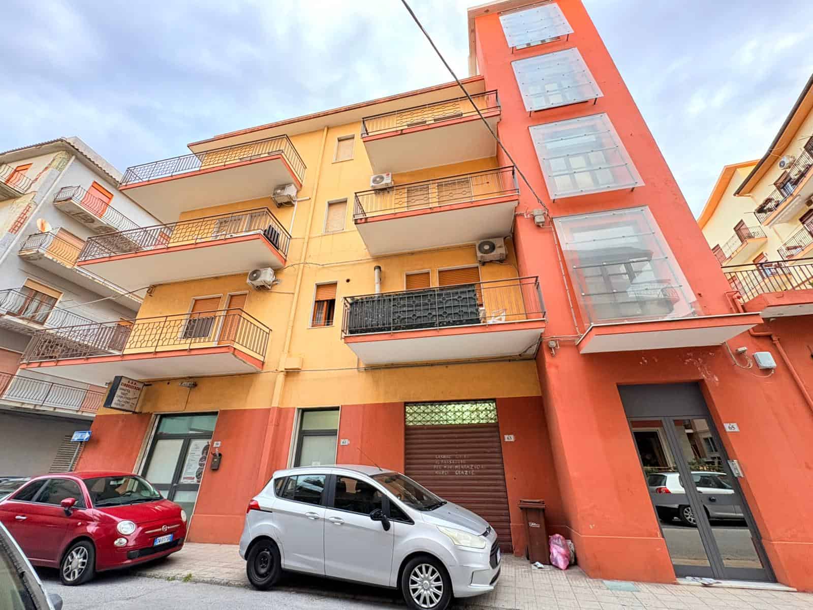 Balcone e facciata di un edificio residenziale colorato con veicoli parcheggiati davanti, in un contesto urbano italiano.