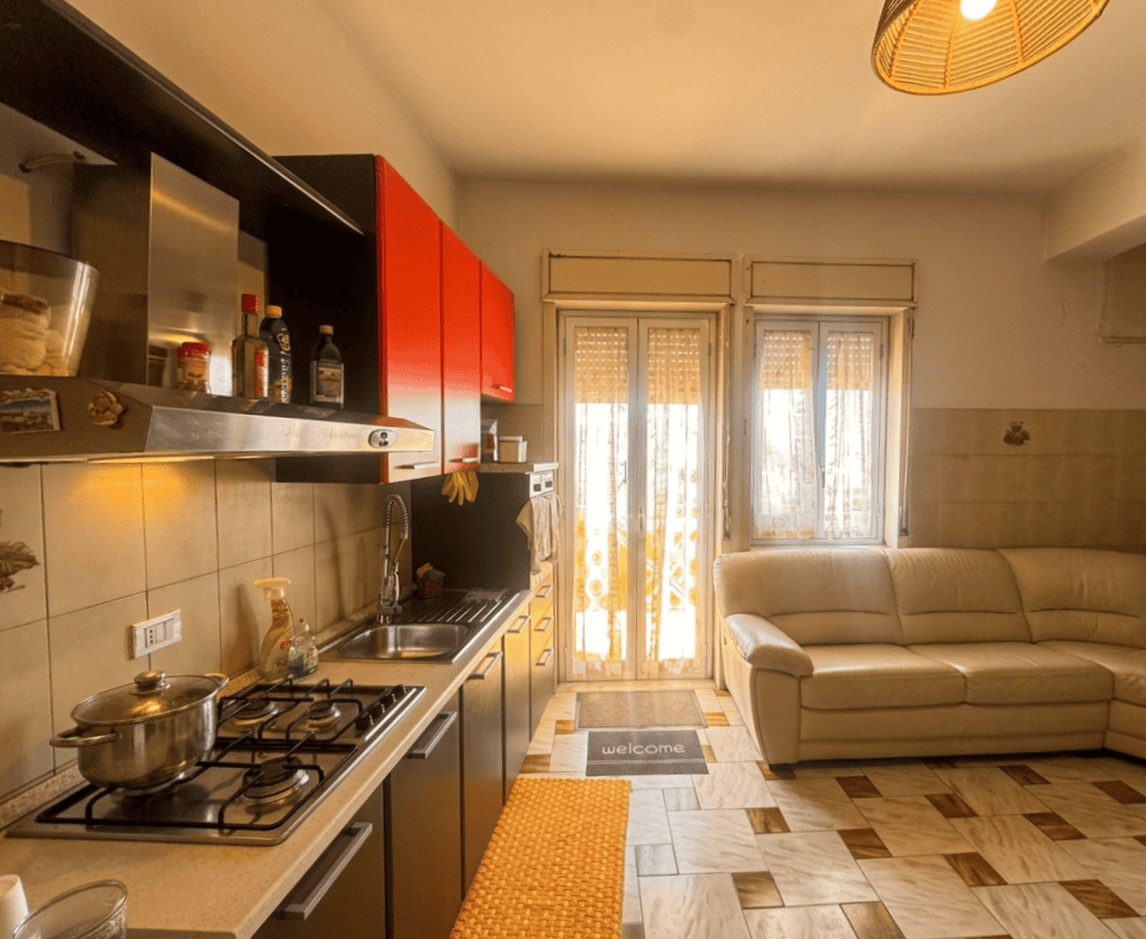 Una cucina moderna e funzionale con mobili rossi e neri, elettrodomestici, e un'area living con divano beige e grandi finestre che lasciano entrare molta luce naturale. Ideale per vivere e ricevere os.