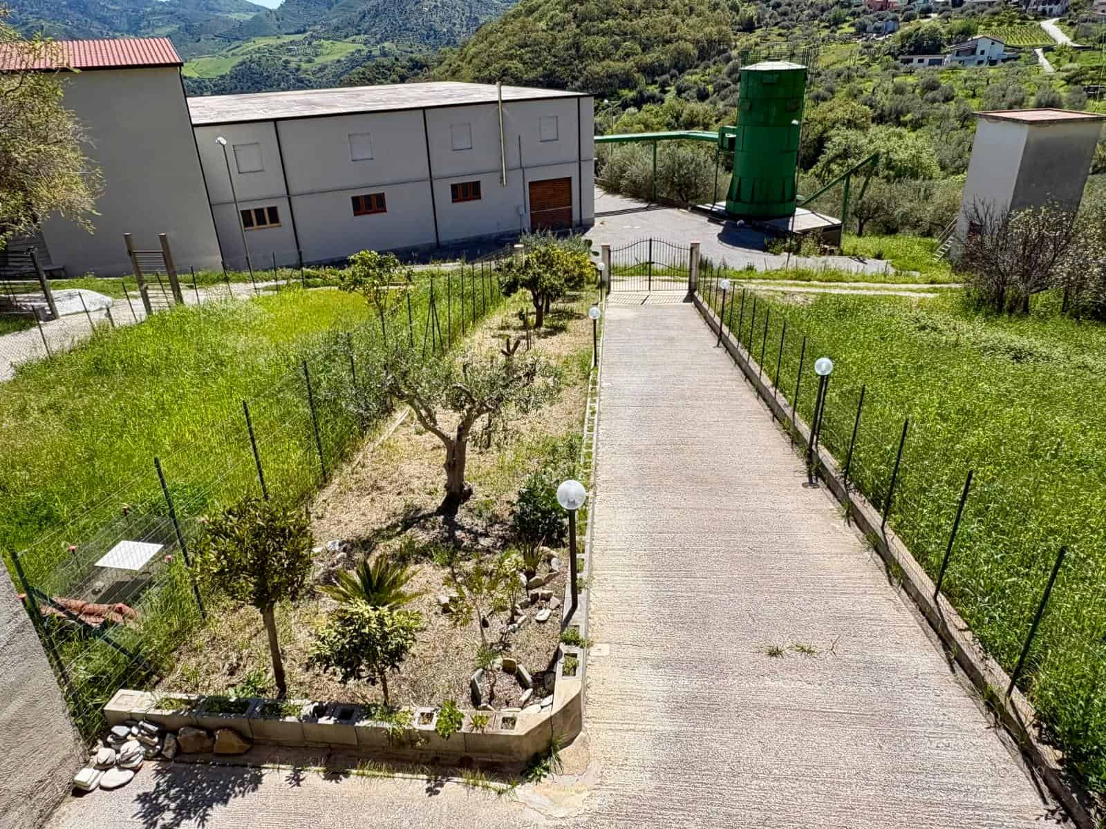 Porte di accesso a un giardino ben curato con piante e alberi, percorso pedonale in cemento e illuminazione esterna, in un'area residenziale tranquilla circondata da natura e montagne.