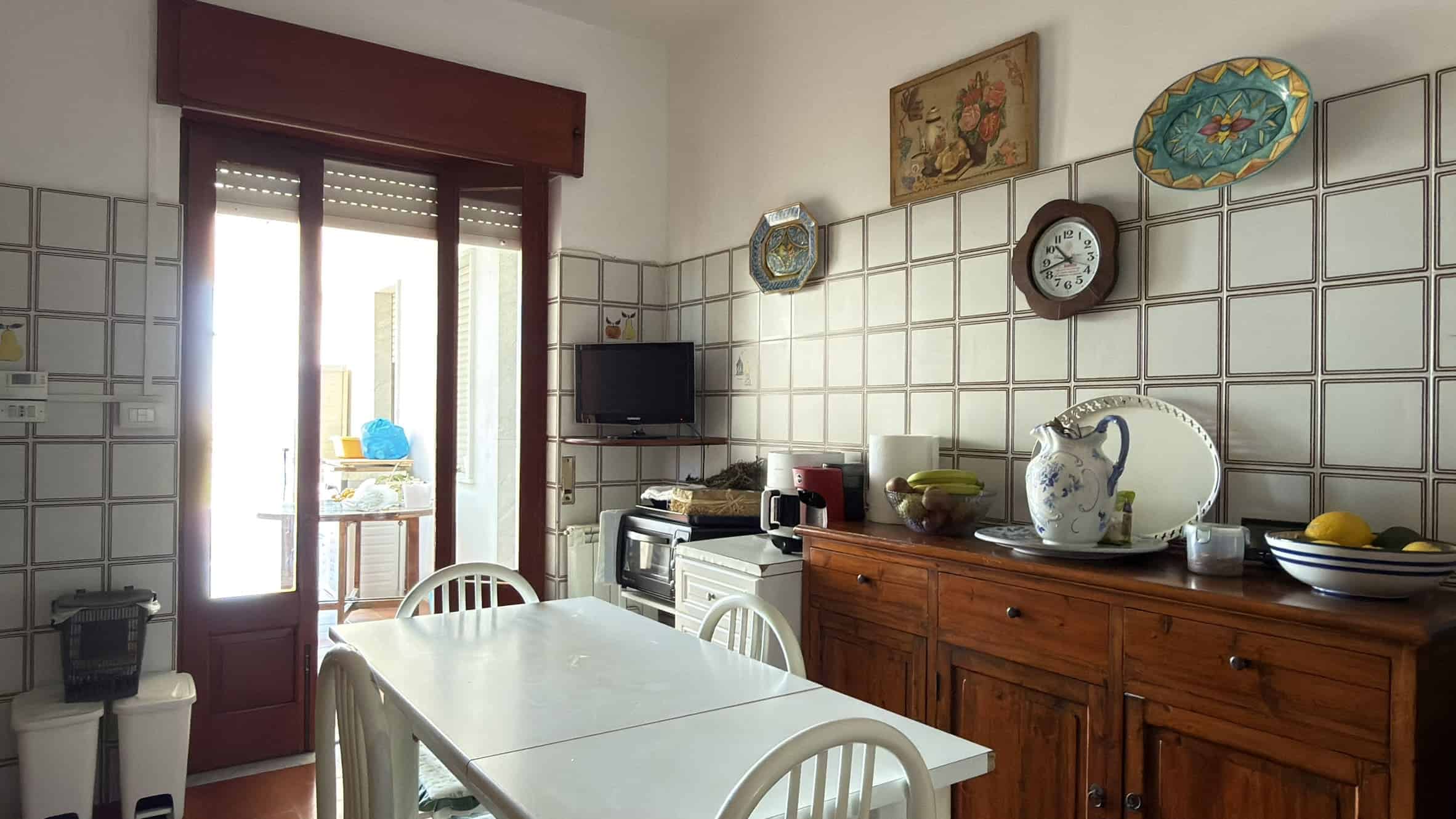 Piatto di cucina con mobili in legno, pareti rivestite di piastrelle bianche, e accessori da cucina come teiera, ciotole e frutta fresca, in un ambiente domestico accogliente e luminoso.