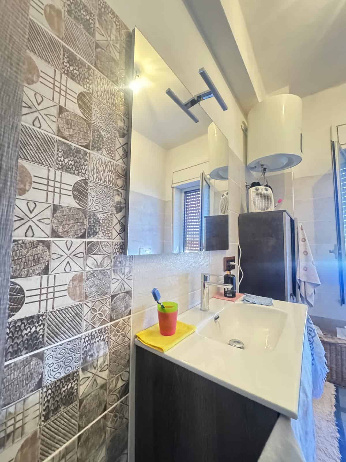 Lavabo moderno in un bagno con pareti rivestite di piastrelle decorative, specchio grande e illuminazione a soffitto, ambiente luminoso e accogliente.