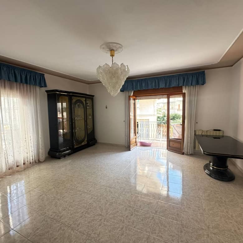 Ampia stanza soggiorno con grandi finestre e balcone, arredamento classico, pavimento in piastrelle lucide, lampadario centrale, pareti decorate con quadri, ideale per vivere in comfort e stile.