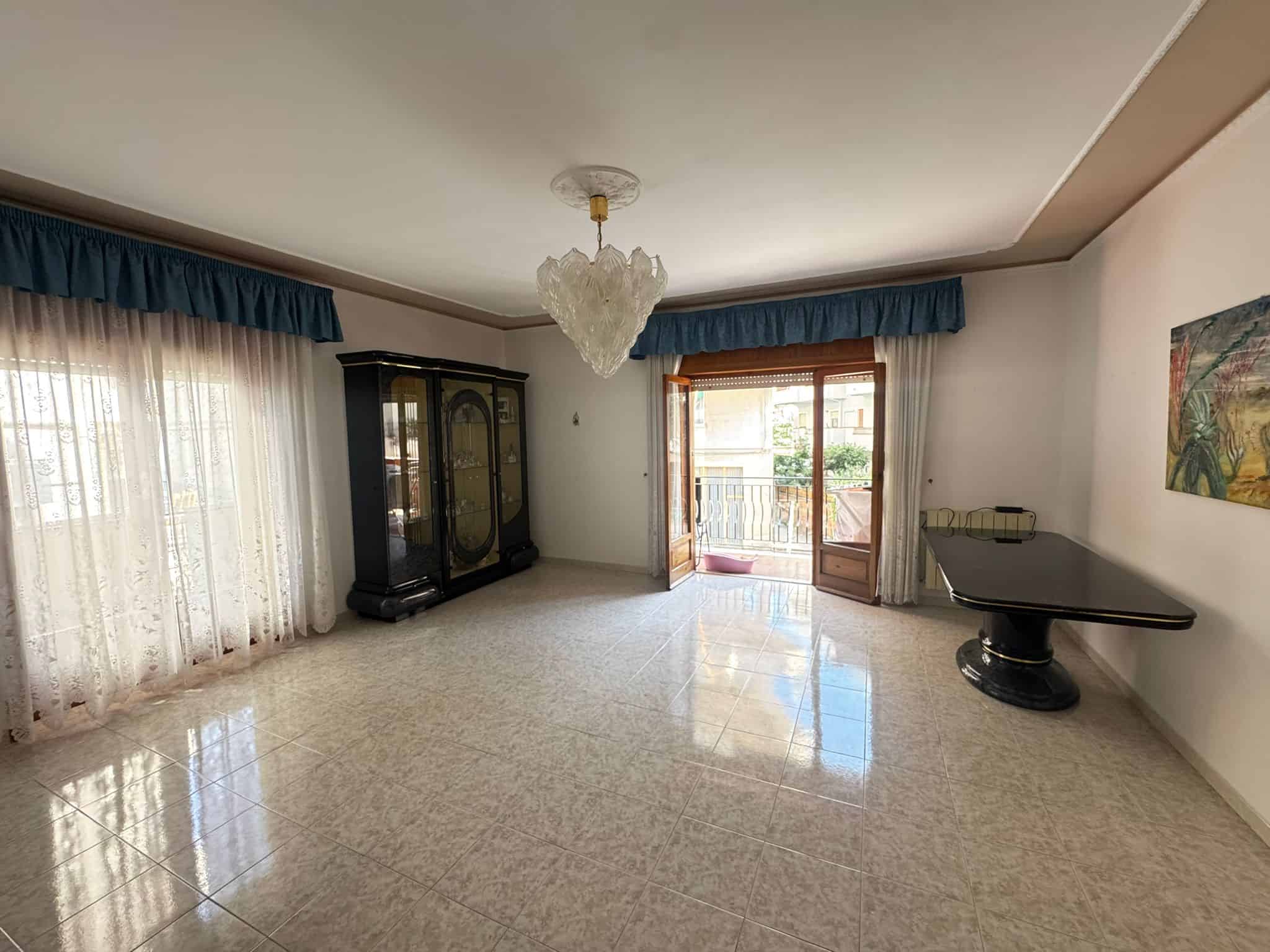 Ampia stanza soggiorno con grandi finestre e balcone, arredamento classico, pavimento in piastrelle lucide, lampadario centrale, pareti decorate con quadri, ideale per vivere in comfort e stile.