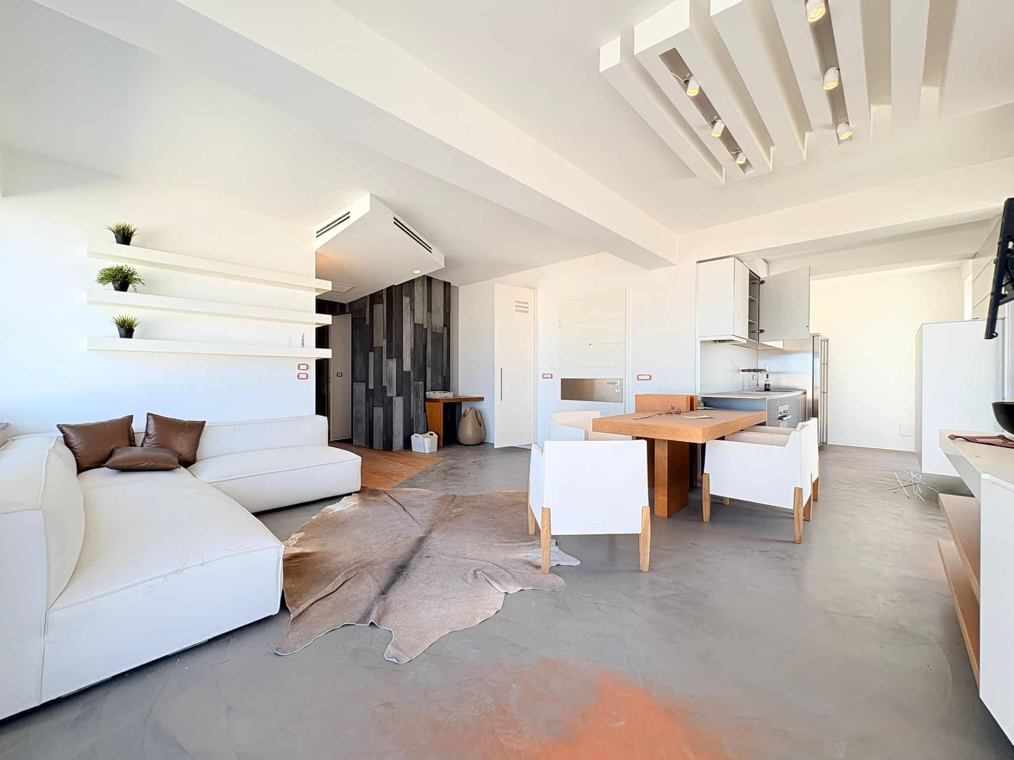 Pannello di soggiorno moderno con divano bianco, tavolo da pranzo in legno e cucina aperta, ambiente luminoso e accogliente con design minimalista e dettagli eleganti.
