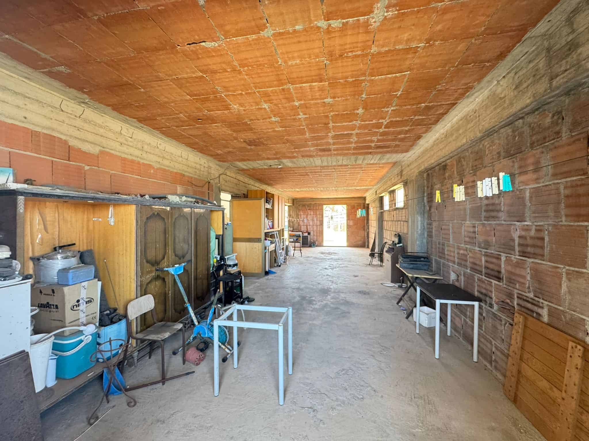 Pannello di costruzione di una casa in fase di ristrutturazione con pareti in mattoni e soffitto in cemento, spazio interno con attrezzi e mobili temporanei, luce naturale dall'ingresso.