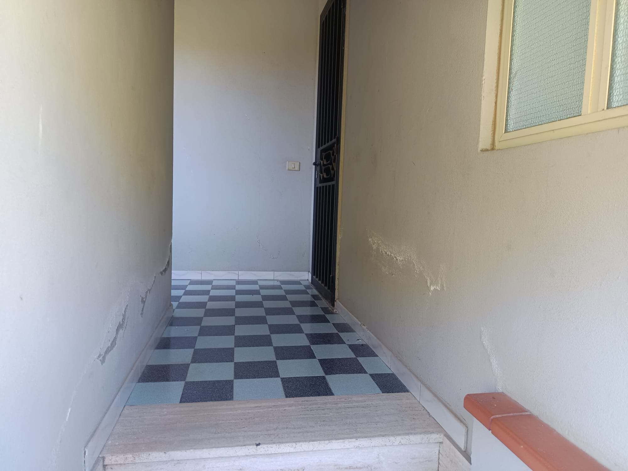 Ampio ingresso esterno di una casa con pavimentazione a scacchi grigi e bianchi, pareti bianche e finestra con vetri opachi, ideale per l'ingresso di un'abitazione.