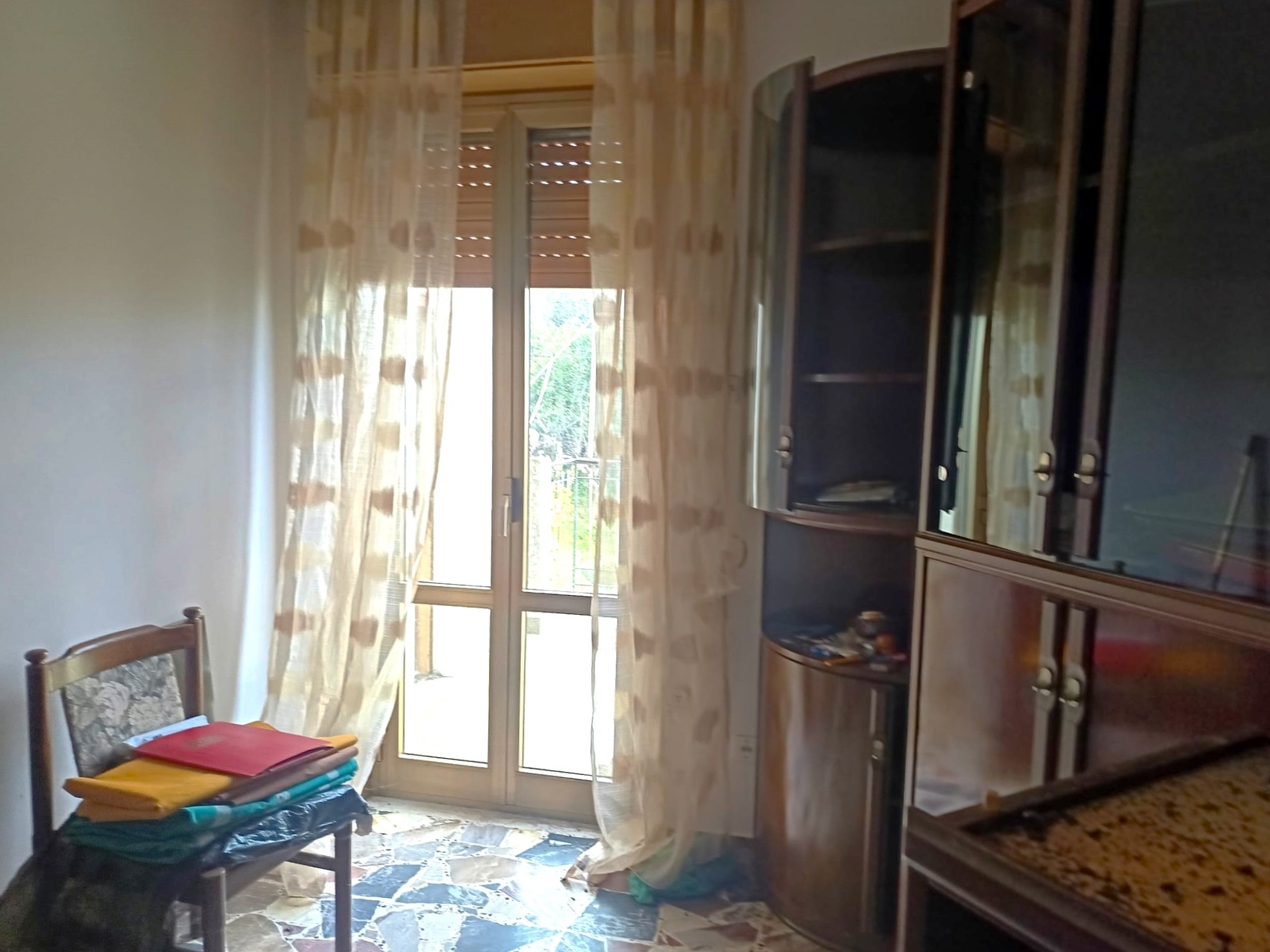Ampia stanza luminosa con porta finestra che dà su un balcone, arredamento in legno scuro, tende leggere con motivo a pois, pavimento in piastrelle di pietra, ideale per ambienti residenziali e relax.