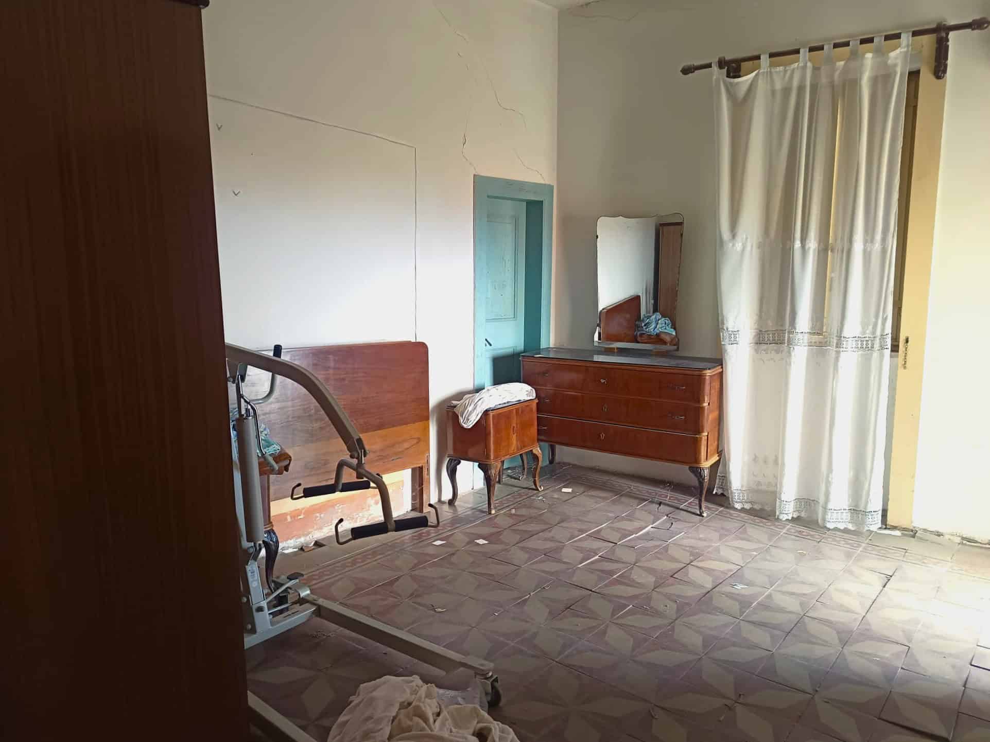 Camera da letto vuota con mobili d'epoca, finestra con tende leggere e pavimento in piastrelle geometriche, ambiente luminoso e semplice.