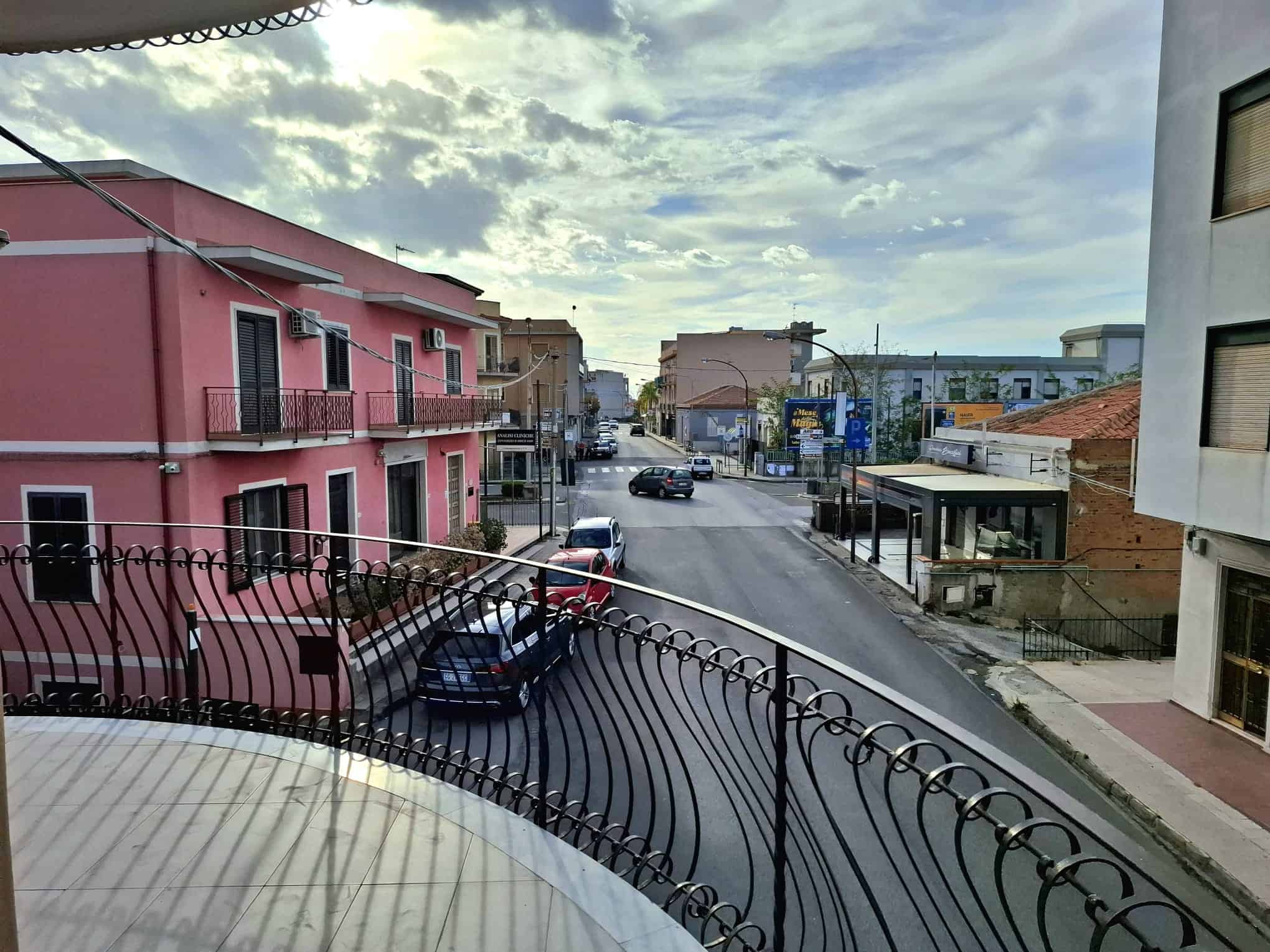 Balcone con vista sulla strada urbana, con edifici colorati e veicoli parcheggiati, sotto un cielo nuvoloso, in un quartiere residenziale di città italiana.