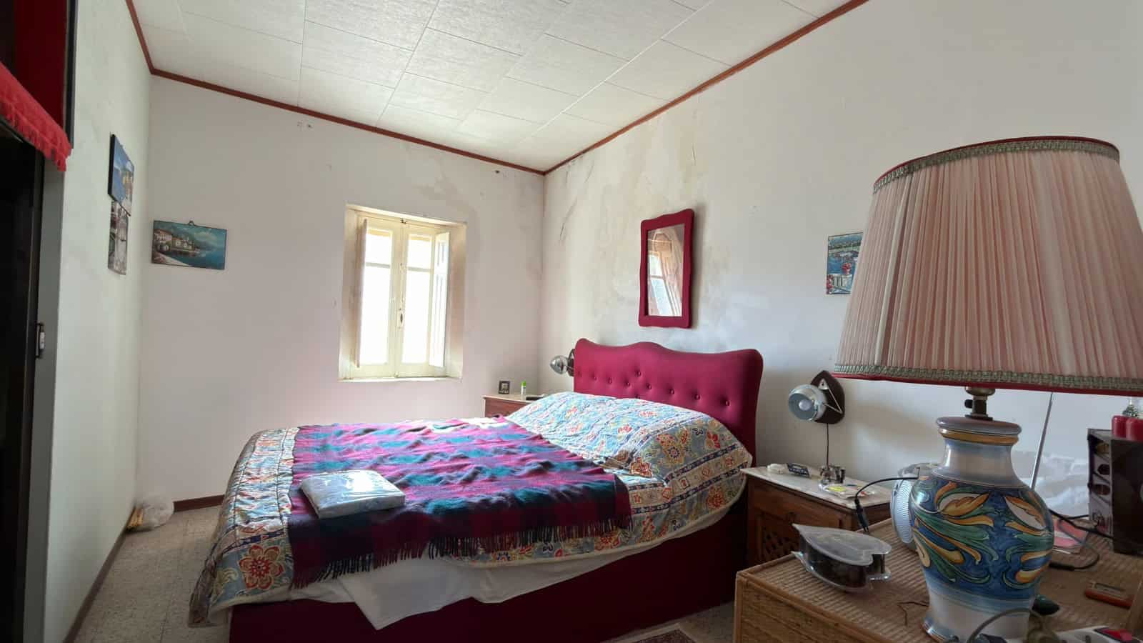 Ampio letto matrimoniale con testiera imbottita rosa, in una stanza semplice e luminosa con pareti bianche, finestra e decorazioni colorate, ideale per affitti a breve termine o soggiorni rilassanti.