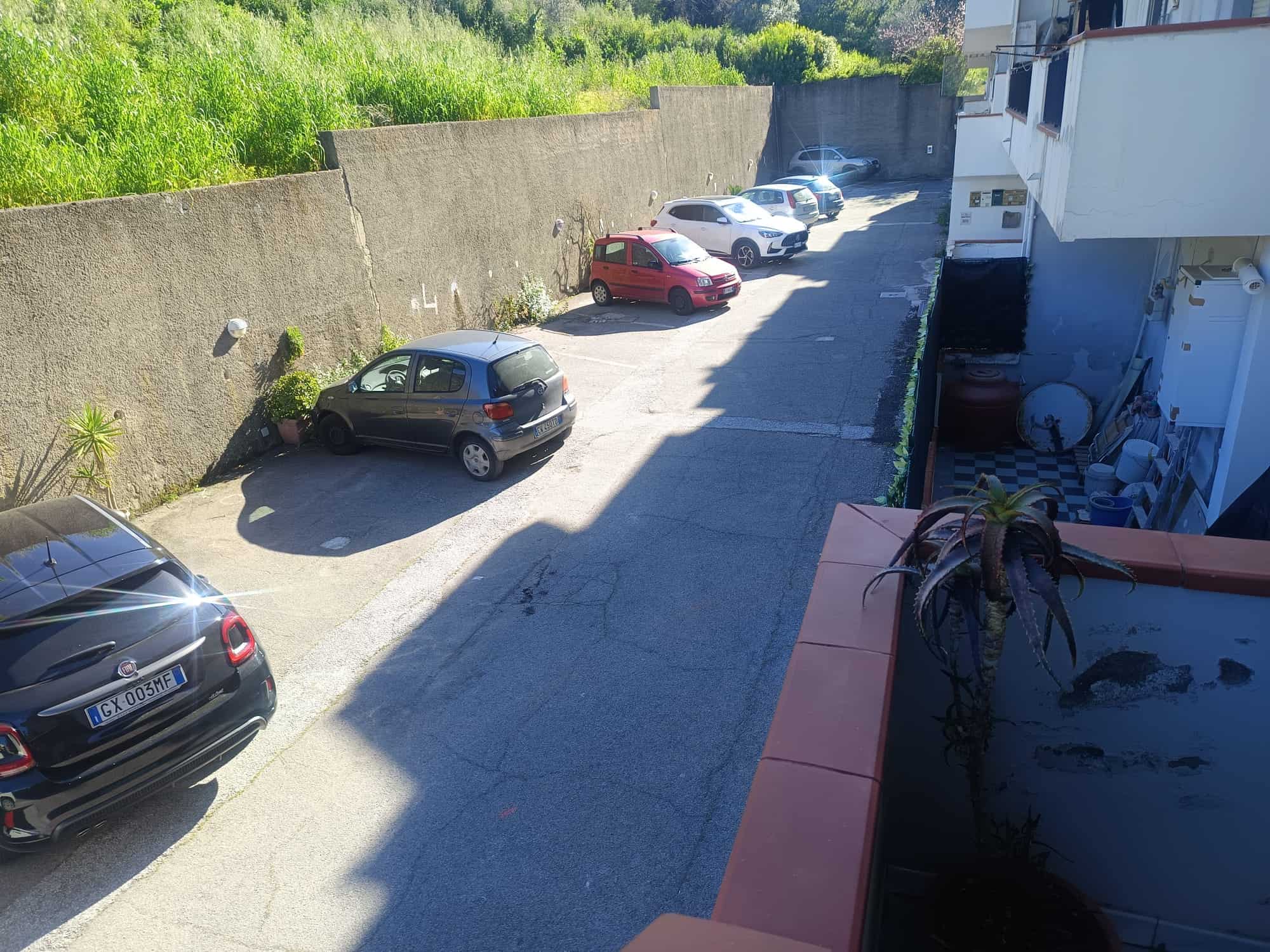 Auto in sosta nel cortile di un edificio residenziale, con parete di cinta in cemento e vegetazione sullo sfondo, in una giornata soleggiata.