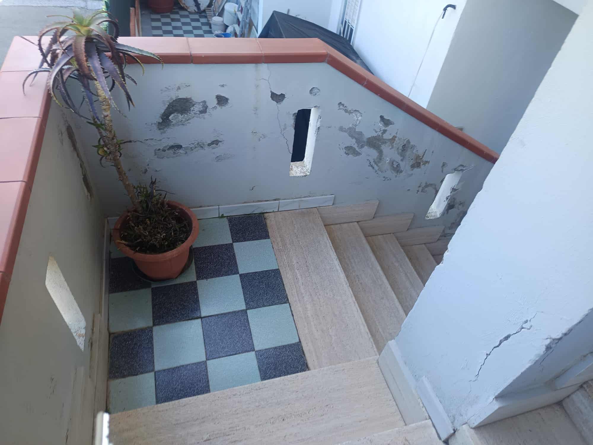 Vedi, un'area esterna di un appartamento con piastrelle a scacchi blu e grigie, una scala in legno e una pianta in vaso, con pareti bianche e una ringhiera in muratura.