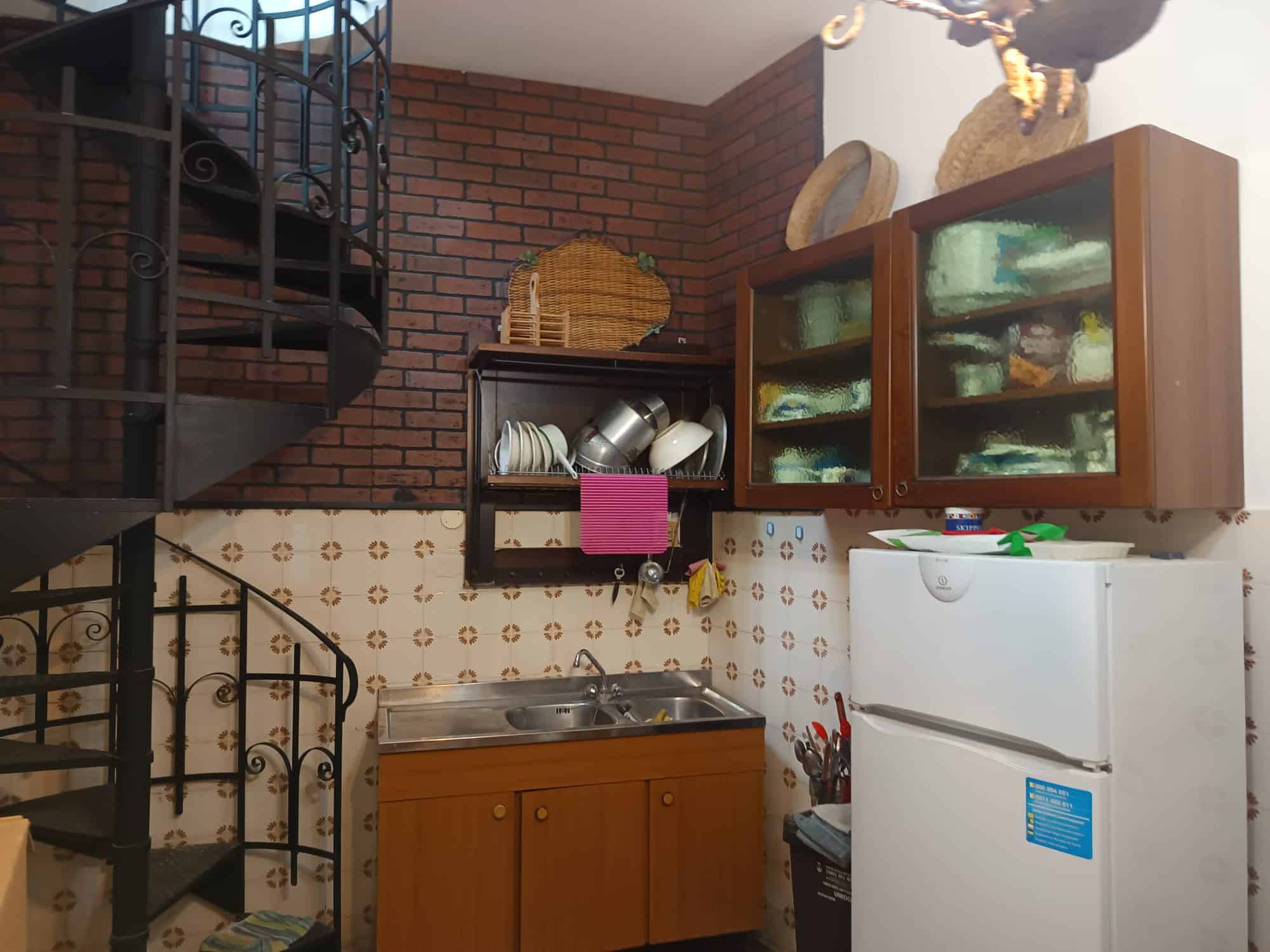Pannello di cucina compatto con armadietti in legno, ripiani aperti e frigorifero bianco in un ambiente domestico con pareti in mattoni e piastrelle decorative.