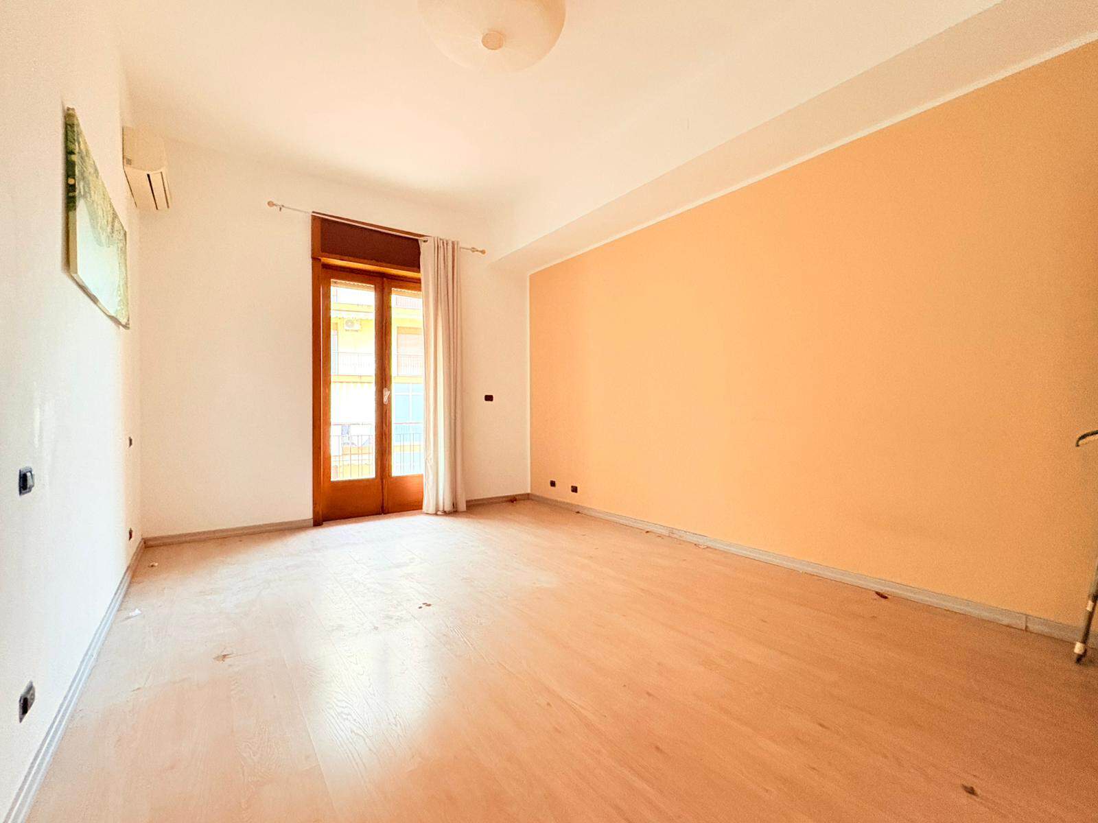 Ampia stanza luminosa con pareti color pesca e pavimento in legno chiaro, ideale per soggiorno o zona living, con accesso a balcone e grandi finestre che lasciano entrare molta luce naturale.