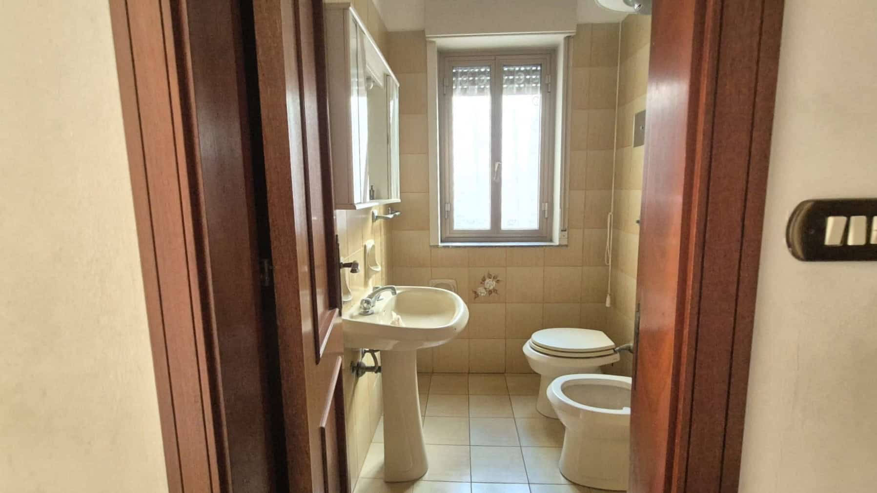Ampio bagno con lavabo, WC e bidet, illuminato da una finestra naturale, con piastrelle beige e arredi in stile classico, ideale per comfort e funzionalità.