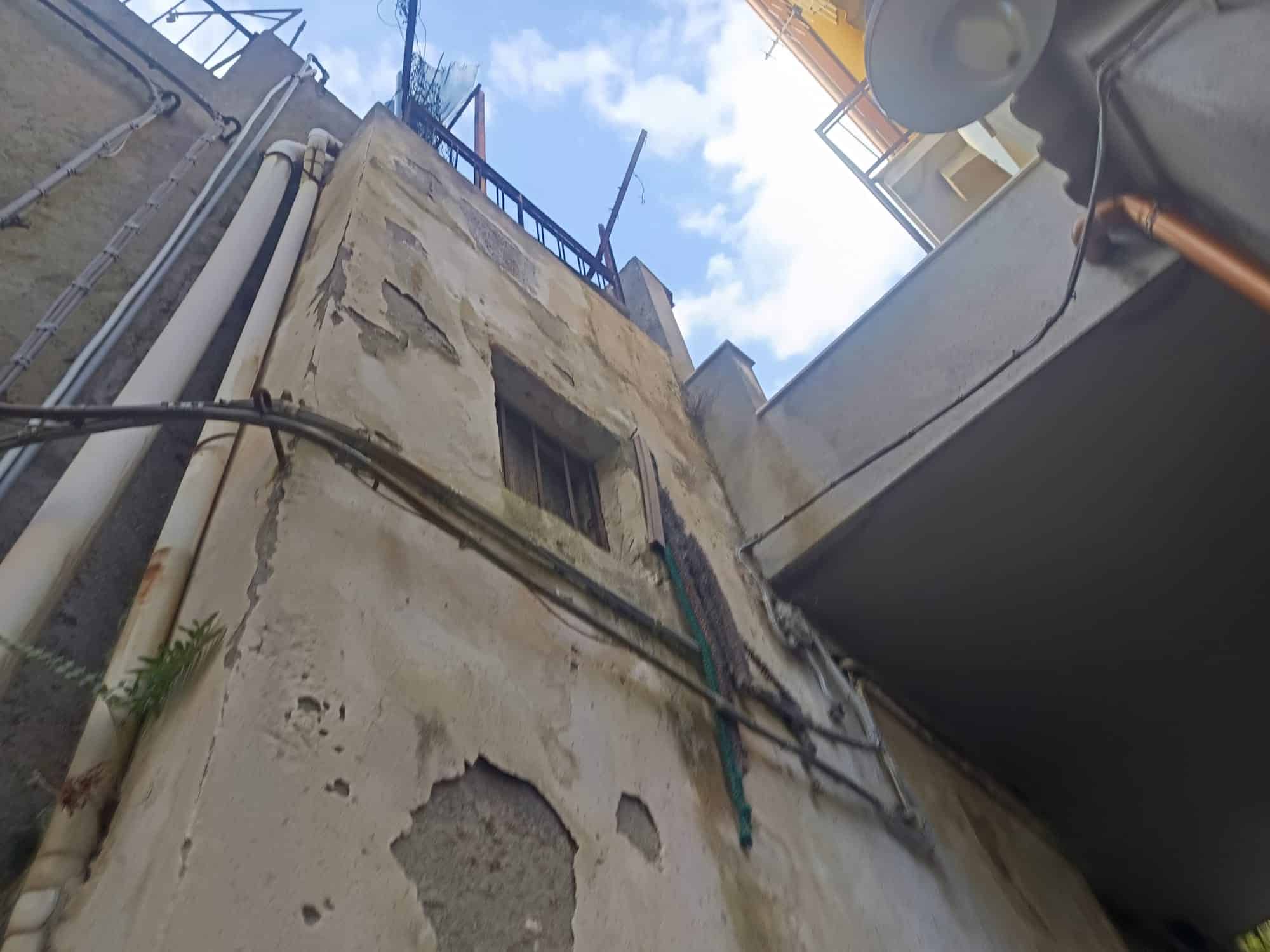 Facciata di un edificio vecchio con pareti danneggiate e tubature visibili, vista dall'angolo con cielo azzurro e nuvole.