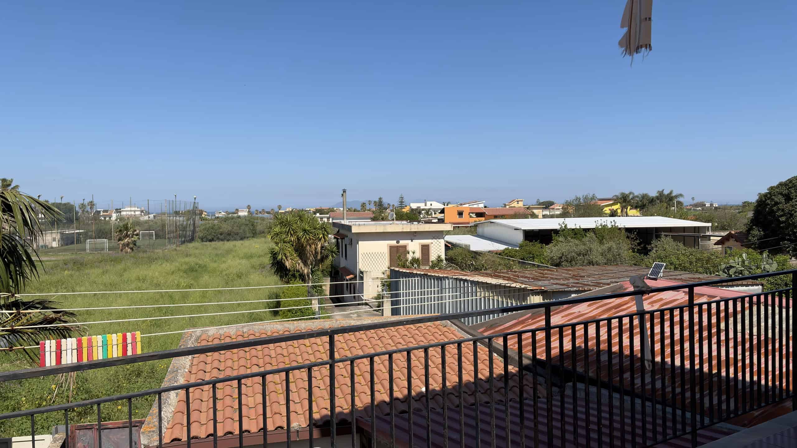 Vista panoramica di un balcone con ringhiera in metallo, che si affaccia su un paesaggio urbano con edifici e spazi verdi, sotto un cielo sereno e azzurro.