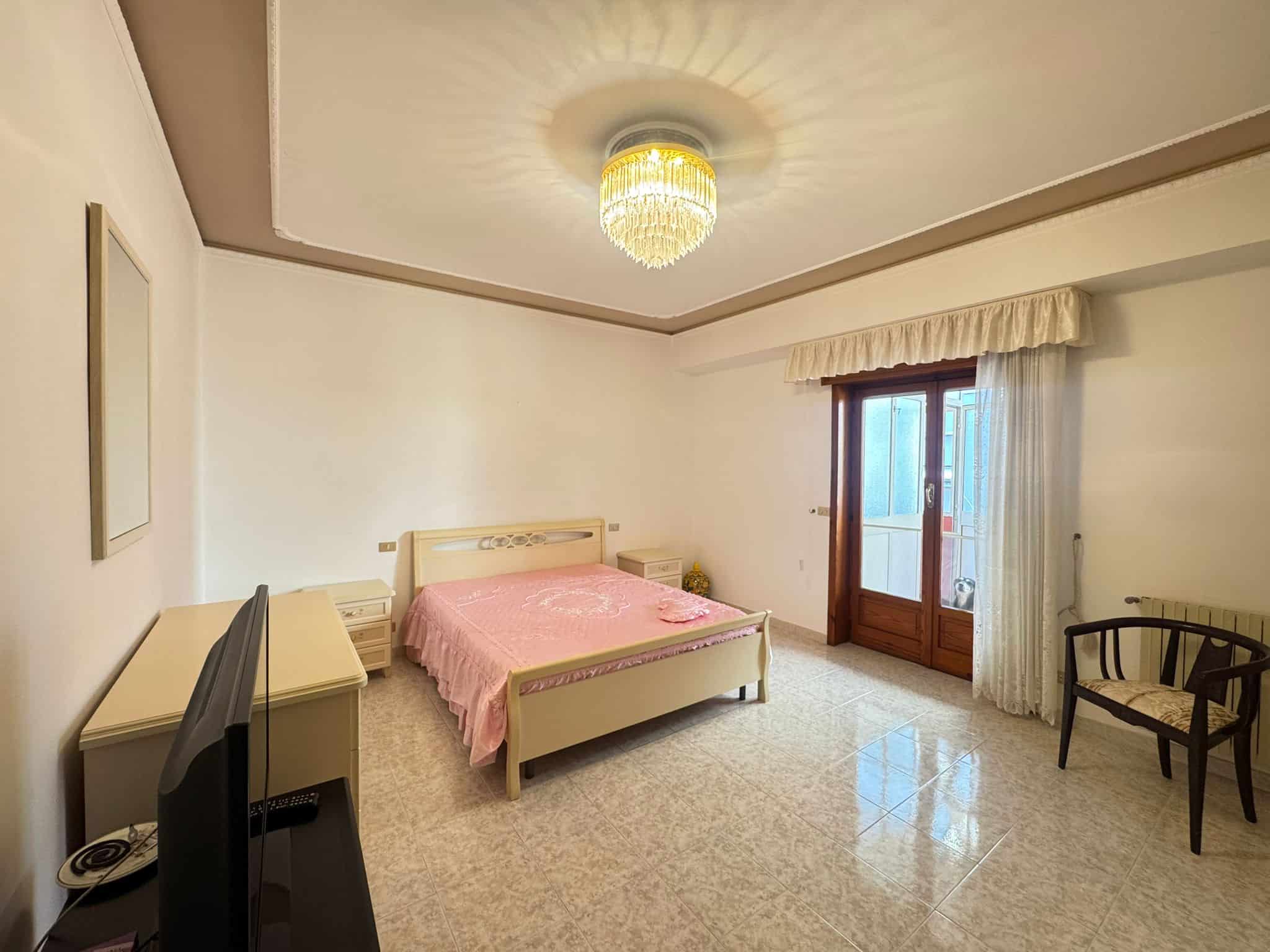 Una stanza da letto luminosa e spaziosa con pareti bianche, letto matrimoniale con copriletto rosa, grande finestra con tende bianche e vista su un balcone, arredamento semplice e elegante, illuminazi.
