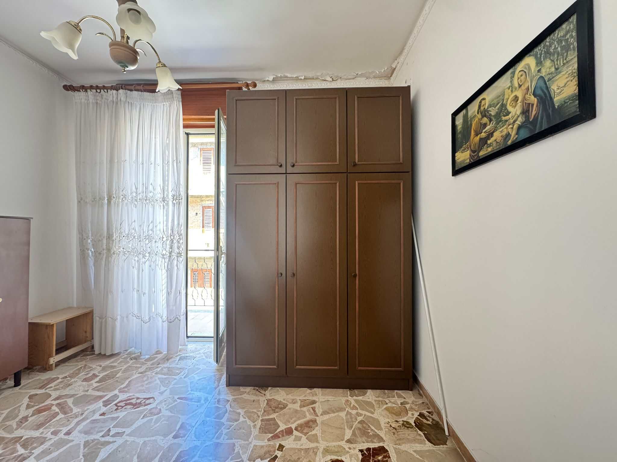 Armadio a due ante in legno scuro in soggiorno con parete bianca, decorato con quadro artistico e illuminato da lampadario a soffitto, vicino a porta finestra con tende leggere.