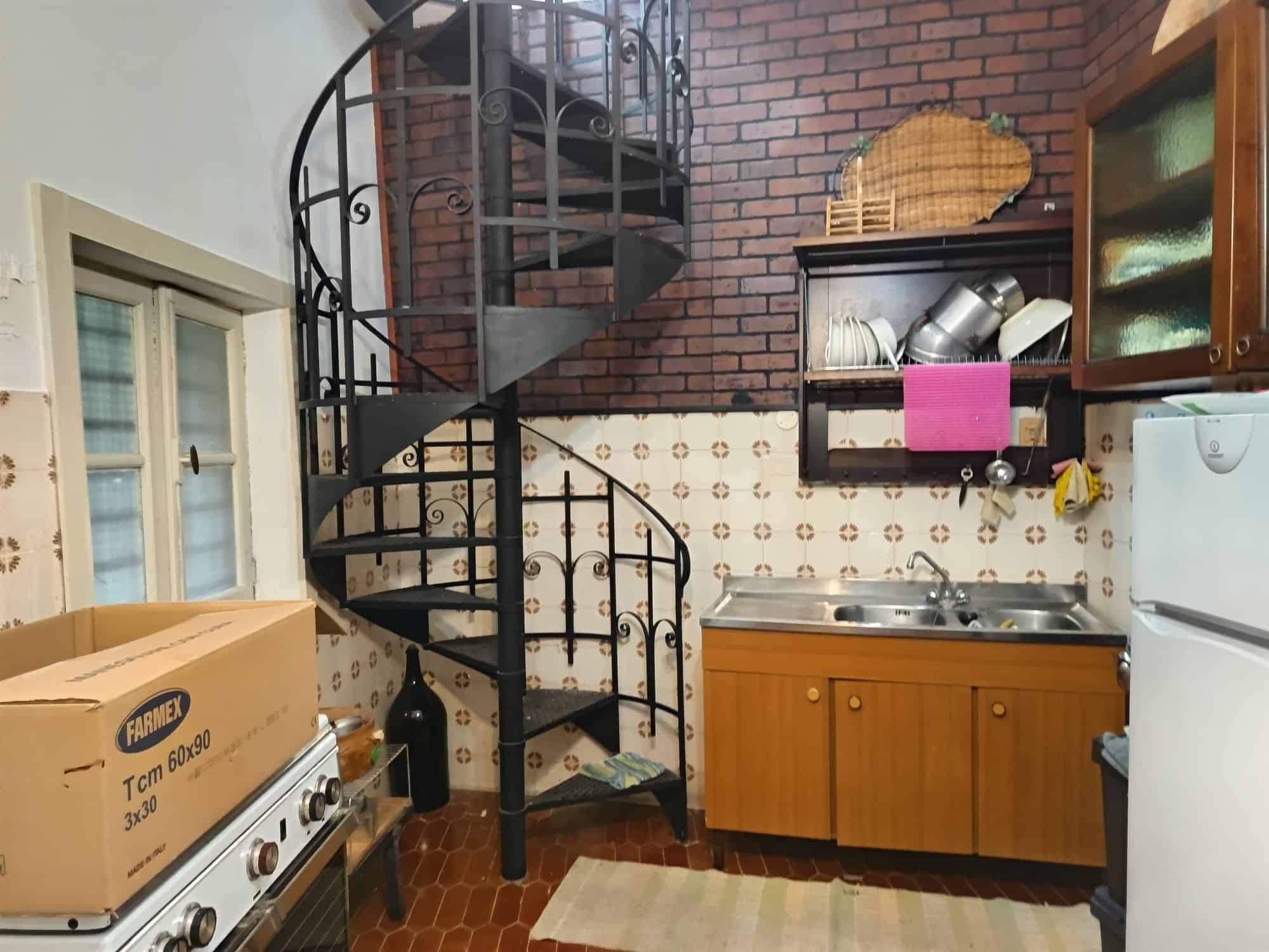Angolo cucina con scala a chiocciola in metallo, mobili in legno, lavello e frigorifero, in un ambiente accogliente e funzionale, ideale per case di charme o appartamenti con spazi ottimizzati.