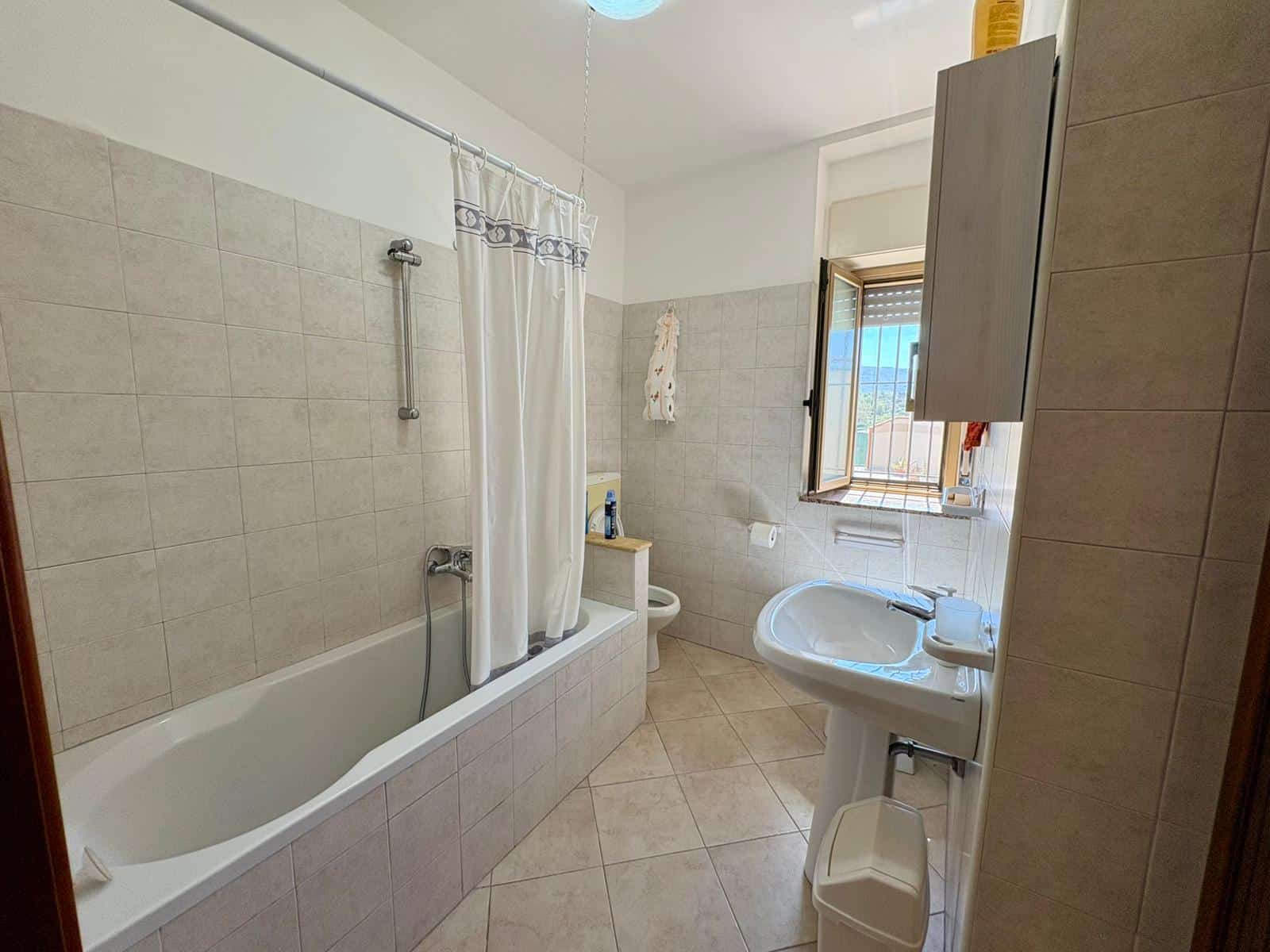 Doccia e vasca da bagno in un bagno spazioso con pareti e pavimenti in piastrelle beige, finestra con vista panoramica, arredi funzionali e illuminazione naturale, ideale per il comfort quotidiano.