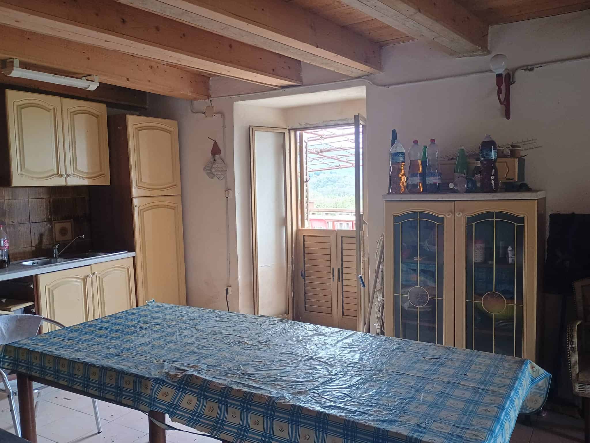 Una stanza abitabile con cucina, tavolo e accessori, illuminata dalla luce naturale proveniente da una porta finestra, con soffitto in legno e arredi in stile rustico.