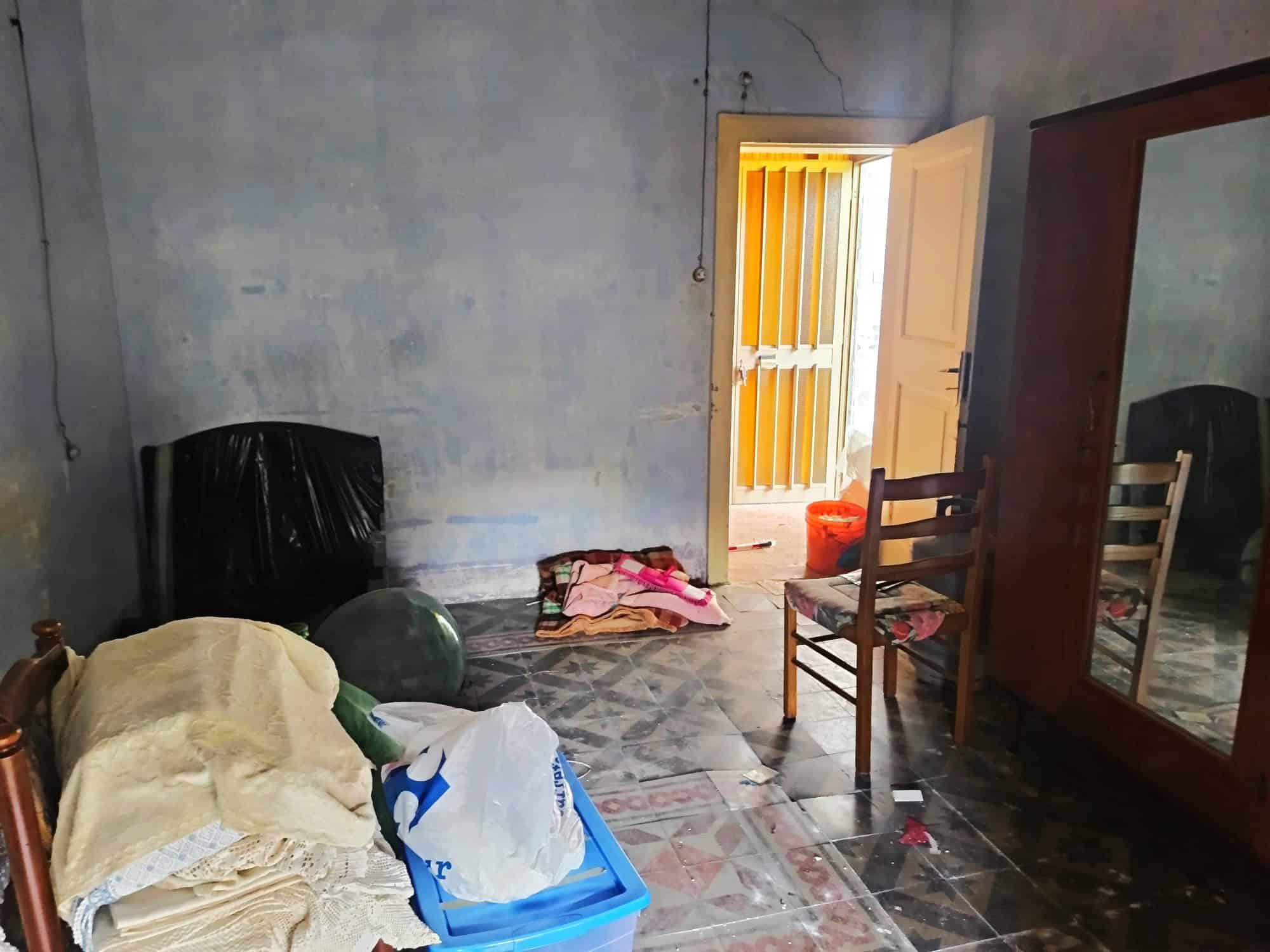 Letto singolo con cuscino e coperta in una stanza con pareti di cemento grezzo, pavimento in piastrelle e porta aperta che conduce a un balcone soleggiato, con mobili e oggetti vari disposti casualmen.