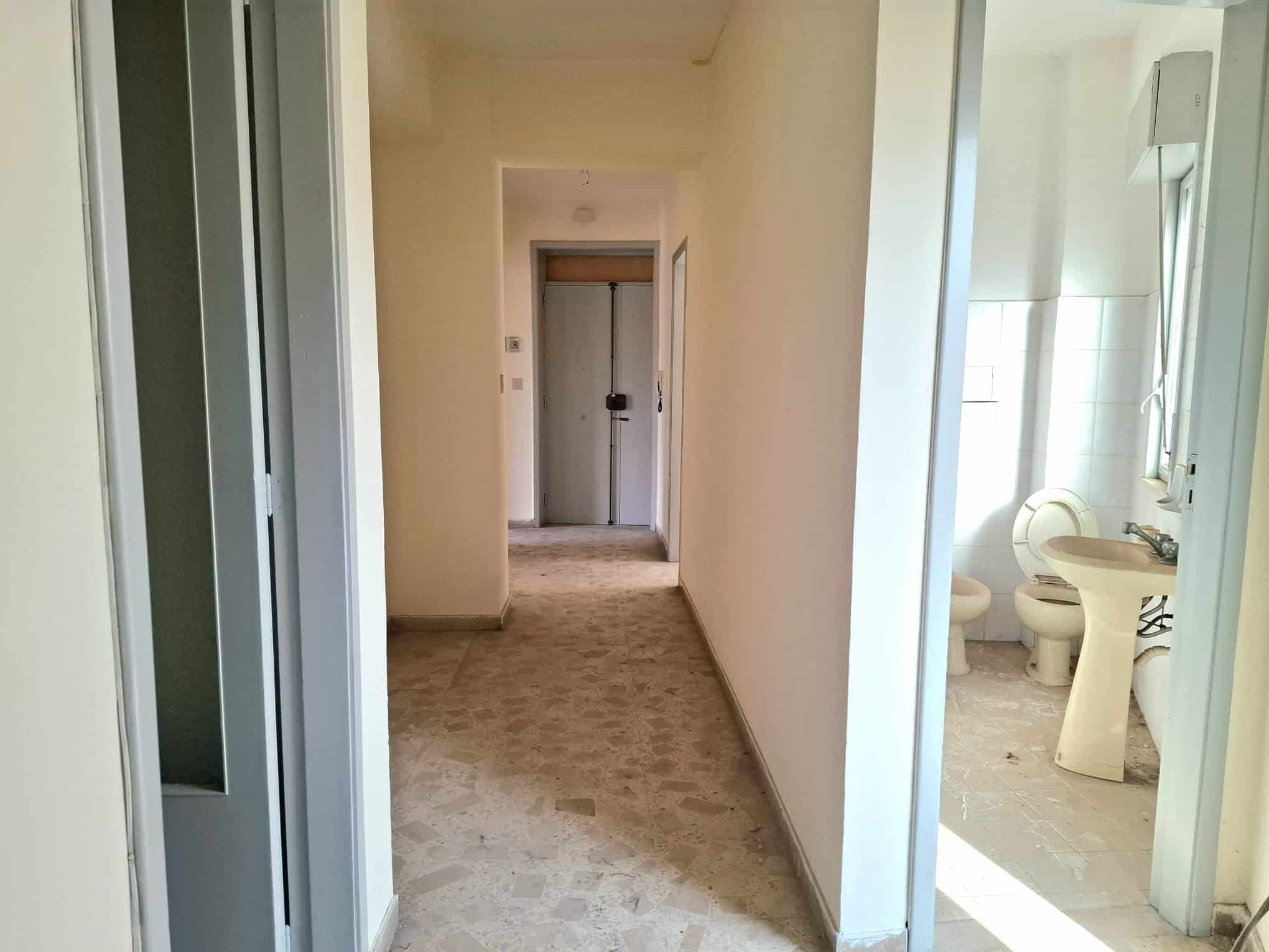 Ampia vista di un corridoio interno di un appartamento con pareti bianche e pavimento in piastrelle beige, che conduce a un ingresso e a un bagno con sanitari visibili.
