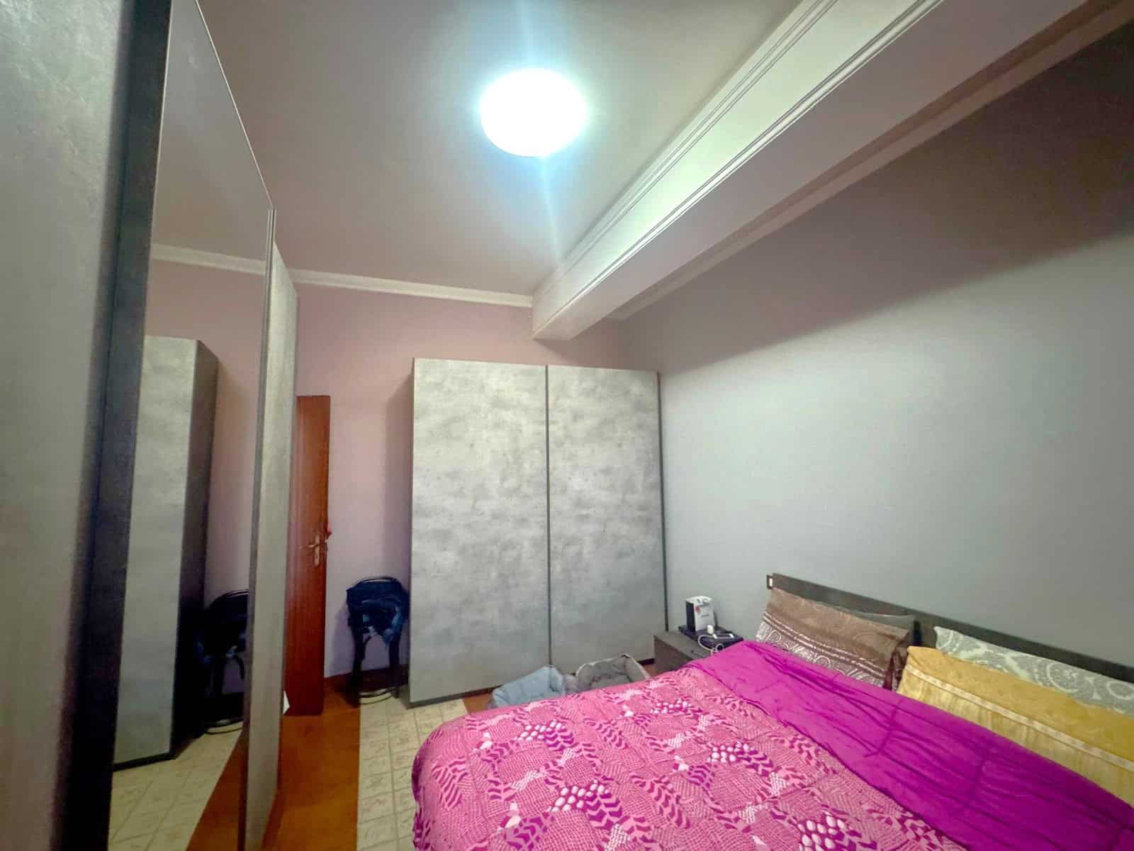 Il letto matrimoniale con copriletto rosa e cuscini decorativi in una camera da letto moderna, con armadio a due ante e pareti color pesca, illuminata da una luce soffusa e un soffitto bianco.