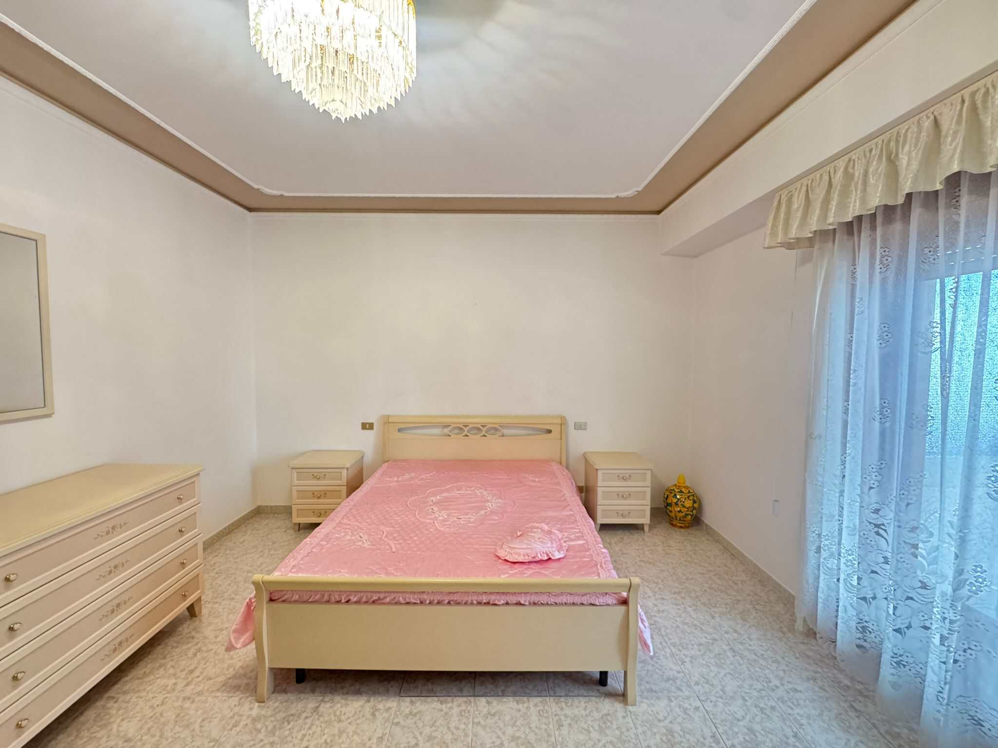 Letto matrimoniale in una camera luminosa con mobili in stile classico, tende leggere e un grande finestrone con vista all'aperto, ideale per case in vendita o affitto.