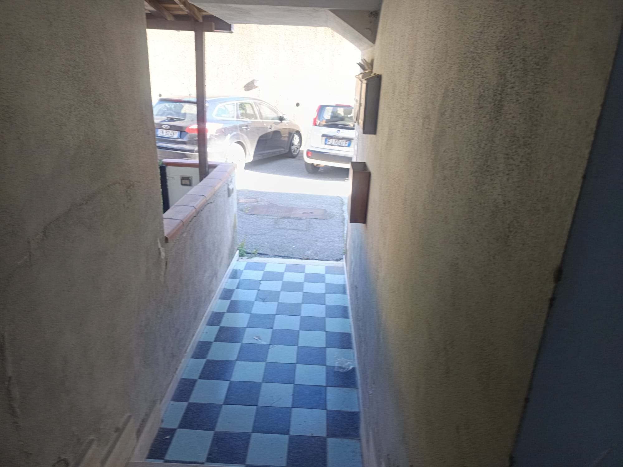 Ingresso di un appartamento con corridoio stretto e pavimento a scacchi blu e bianco, con vista verso il parcheggio esterno e le auto parcheggiate.