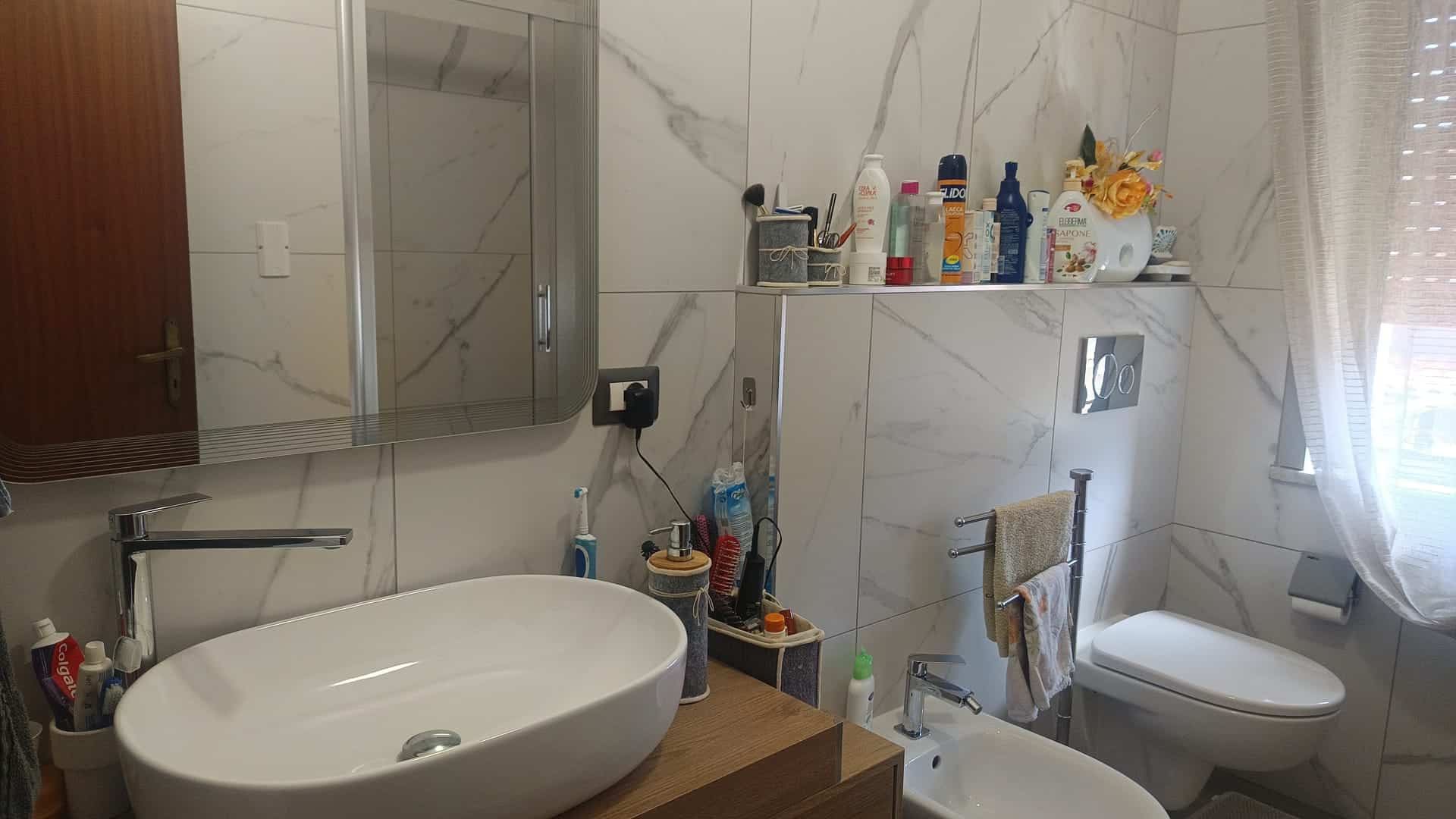 Lavabo moderno in bagno con pareti in marmo bianco, accessori e prodotti per l'igiene, finestra con tende leggere, ambiente luminoso e elegante.