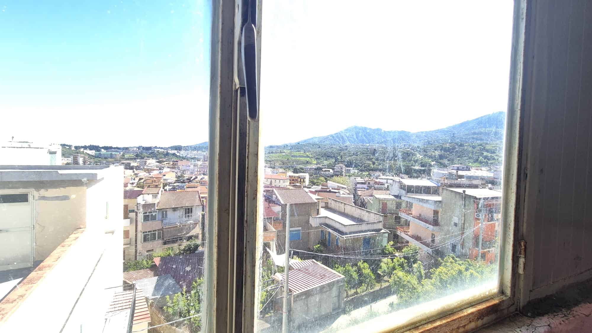 Vista panoramica della città e delle montagne dal finestrino di un appartamento, con paesaggio urbano e natura visibili attraverso il vetro.