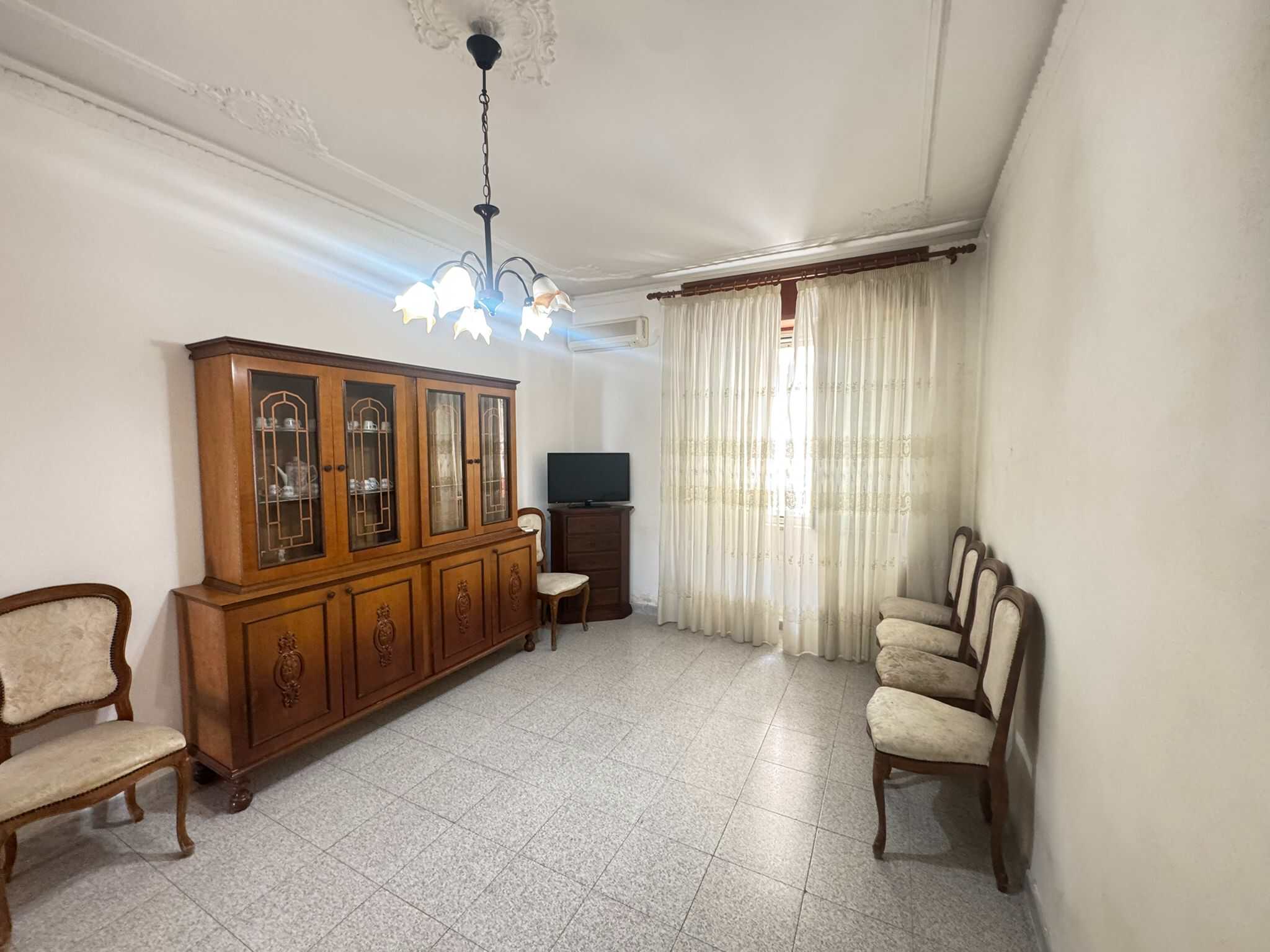 Sala da pranzo spaziosa con mobili classici, grande finestra con tende leggere e illuminazione centrale, ideale per soggiorni confortevoli.