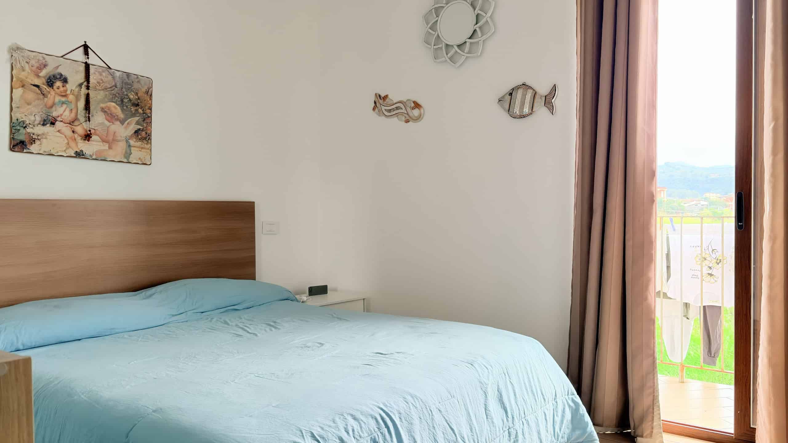 Ampia camera da letto con letto matrimoniale, pareti decorate con quadri e pesci in metallo, grande finestra con vista panoramica e tende beige, ambiente luminoso e accogliente.