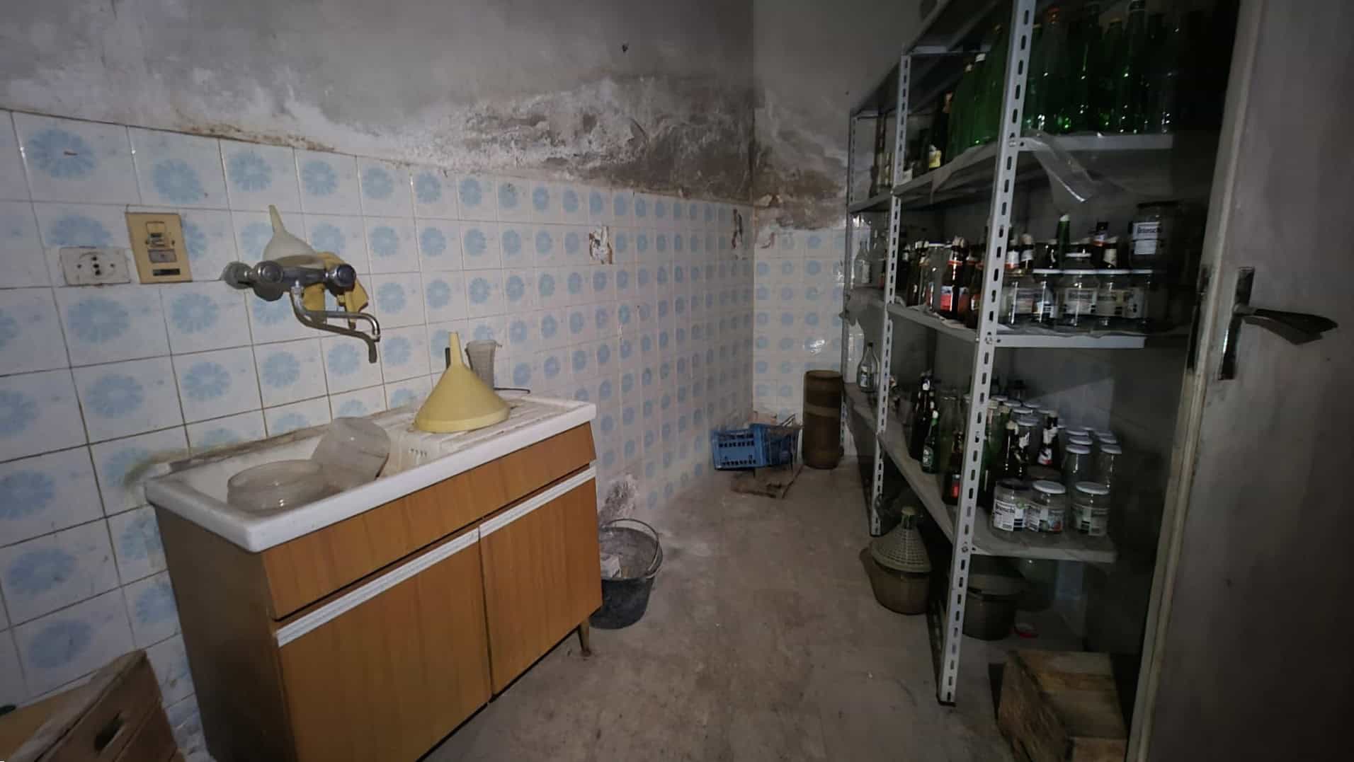 Lavandino in un locale di servizio con scaffali metallici pieni di bottiglie e contenitori, pareti in mattoni e piastrelle, ambiente funzionale e pratico per le faccende domestiche.