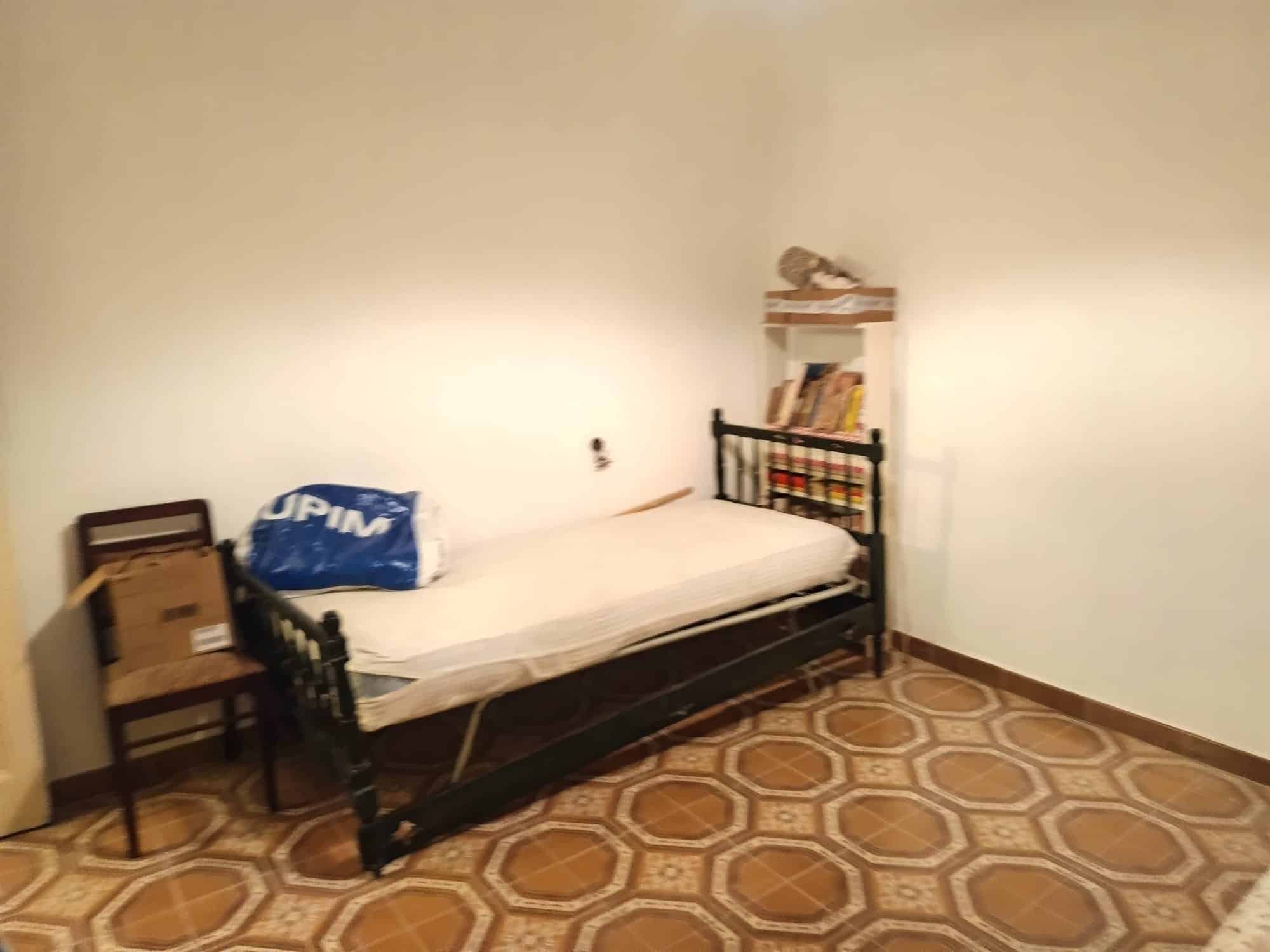 Letto singolo con struttura in metallo nero, in una stanza semplice con pareti bianche e pavimento in piastrelle geometriche. Accanto al letto, una sedia in legno con alcuni oggetti e una piccola mens.
