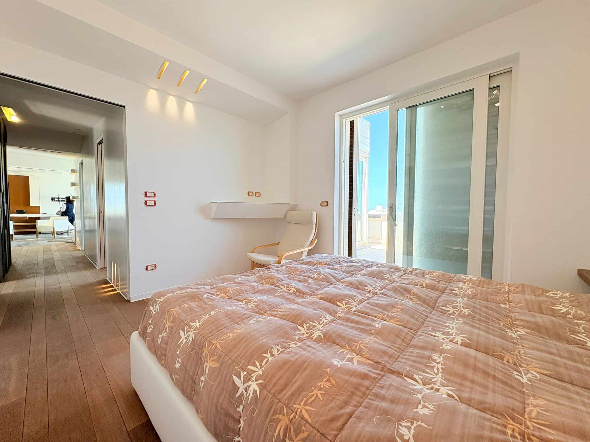 Appartamento moderno con camera da letto luminosa e balcone con vista sul mare, arredamento elegante e pavimento in legno, ideale per soggiorni di relax e comfort.