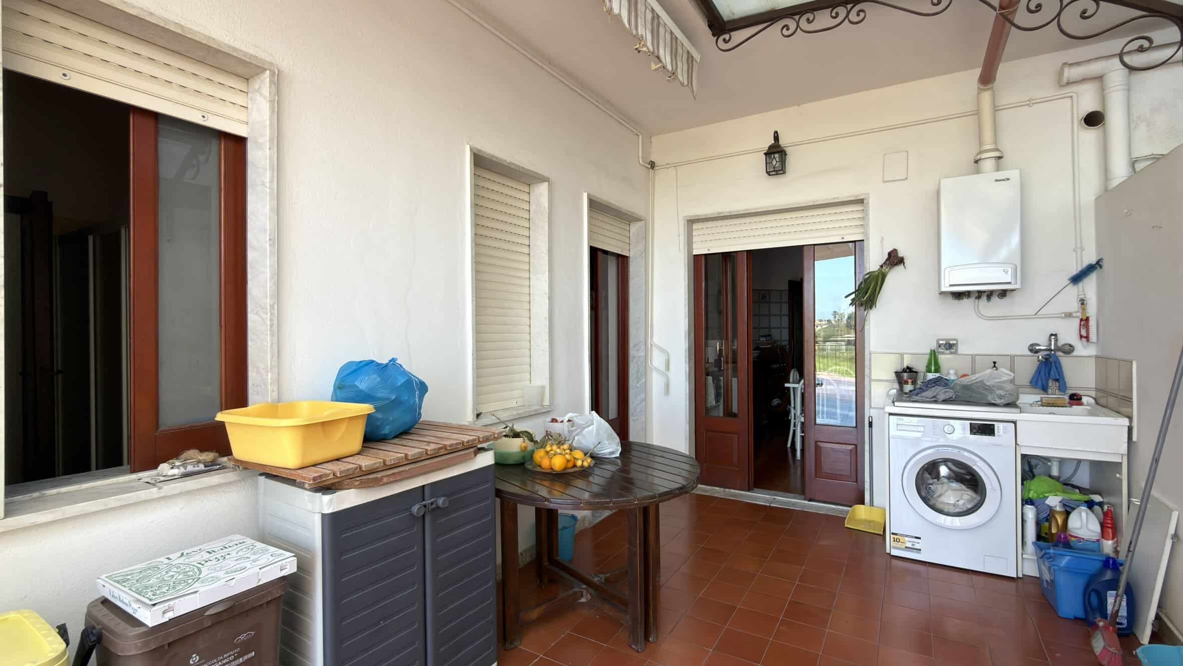 Lavanderia esterna con lavatrice, lavandino e spazio di stoccaggio, ambiente pratico e funzionale per le faccende domestiche all'aperto.