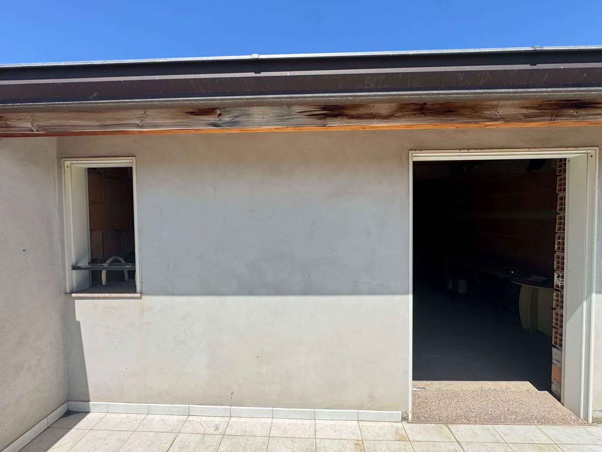 Finestra e porta garage su parete esterna di una casa moderna, con pareti bianche e pavimento in piastrelle, sotto un cielo azzurro.