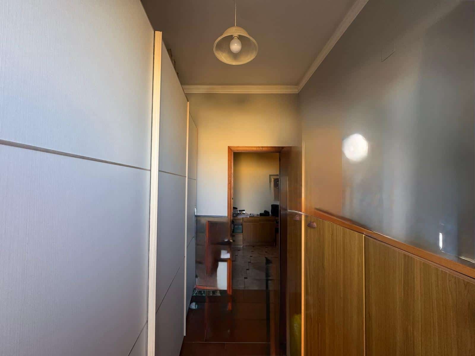 L'immagine mostra un corridoio interno di un appartamento con pareti chiare e pavimento in legno, illuminato da una luce centrale e da una lampada a soffitto, con una porta aperta che conduce a un'alt.