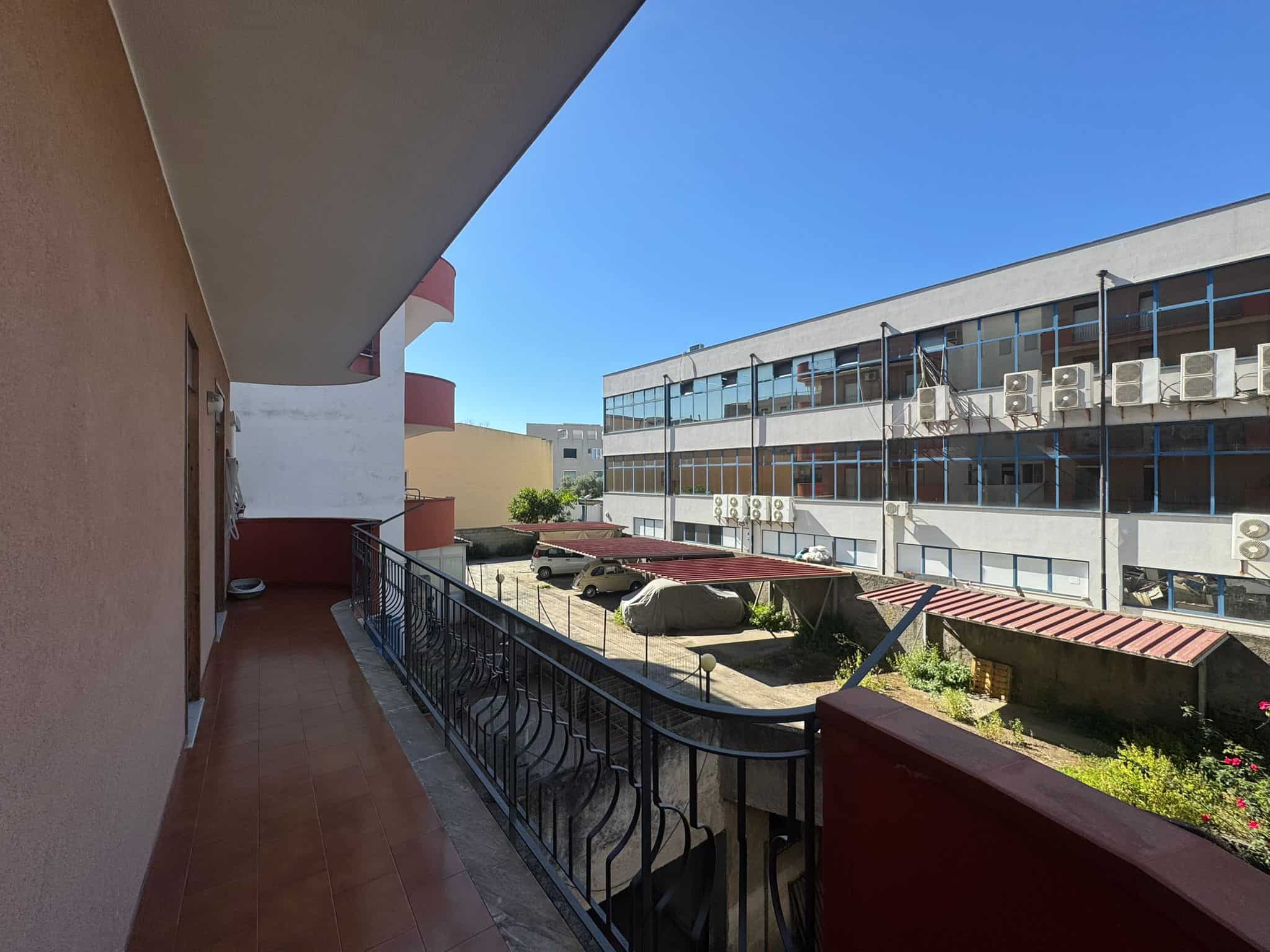 Balcone di un appartamento con vista su edifici commerciali e aree verdi, in un complesso urbano moderno, con un cielo sereno e aria fresca, ideale per relax e momenti di tranquillità.