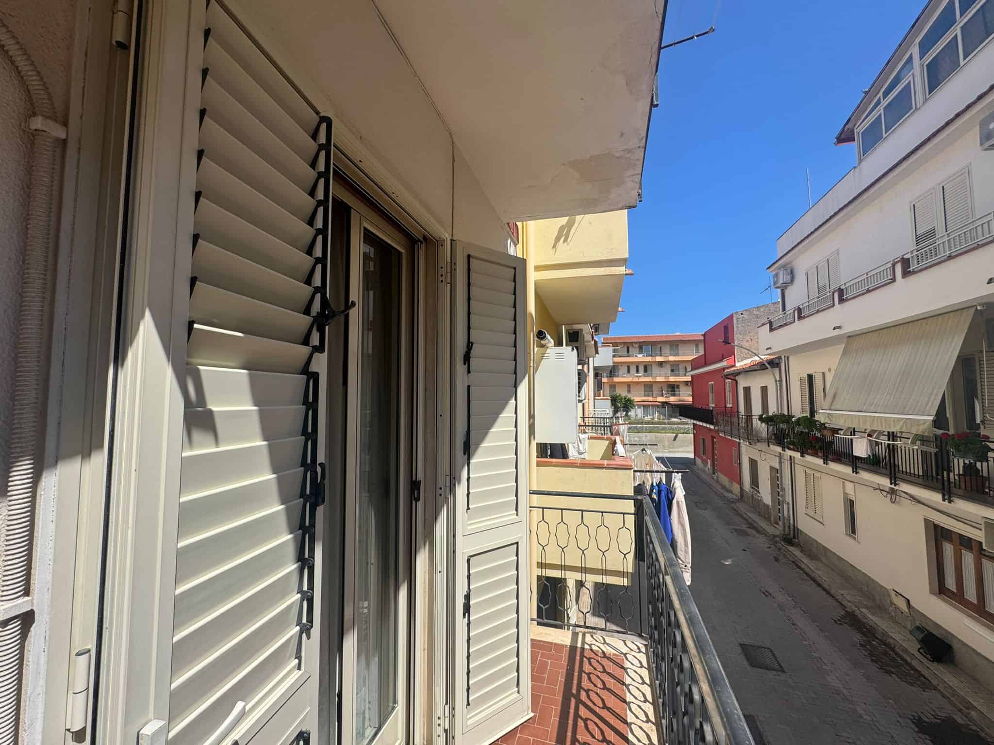 Ampio balcone con persiane in alluminio e vista su strada residenziale, ideale per relax e momenti all'aperto in un contesto urbano tranquillo.