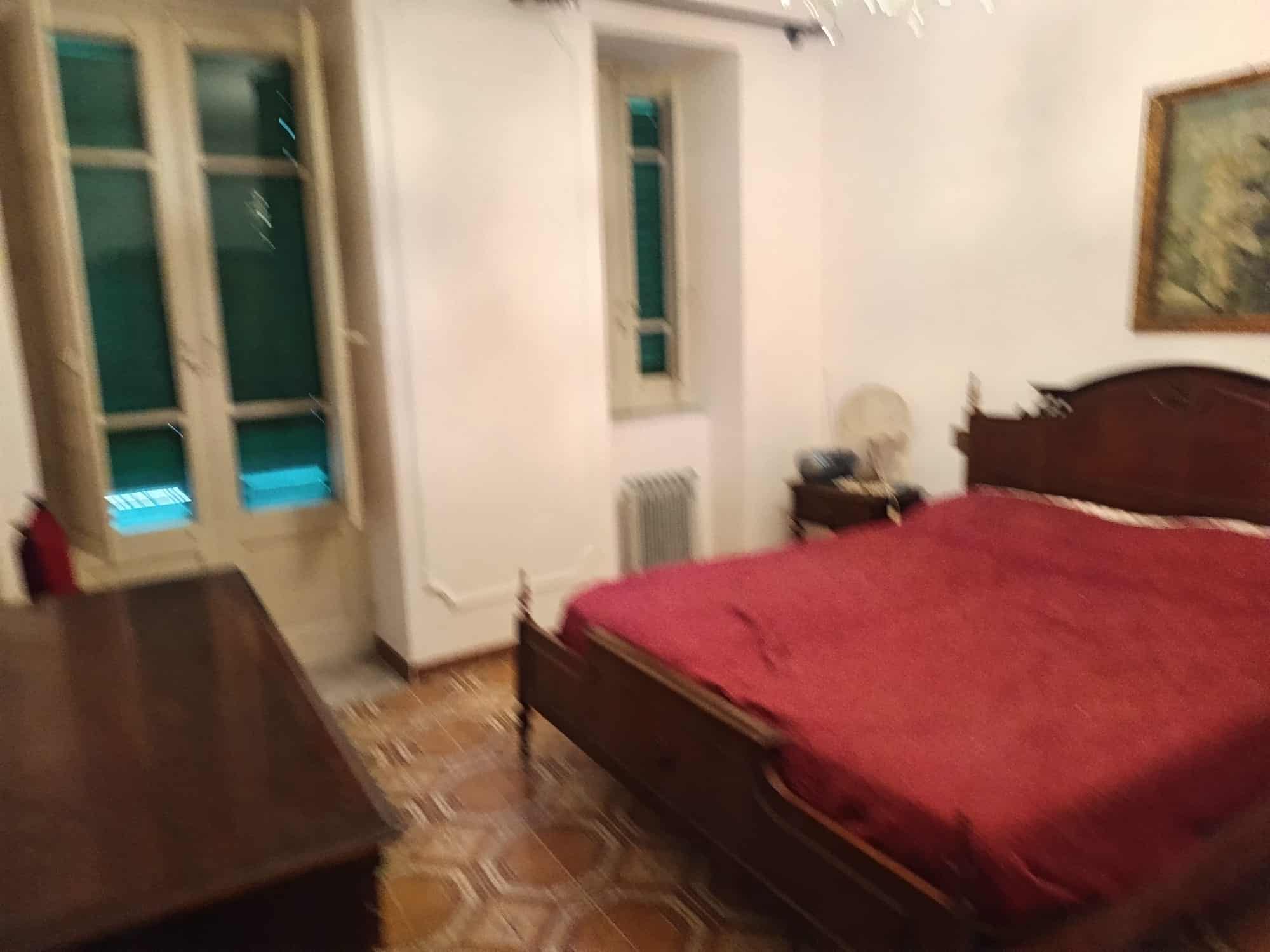 Una stanza da letto con letto matrimoniale in legno, lenzuola rosse, e arredi classici, con pareti bianche, finestra e quadro decorativo, in un ambiente accogliente e elegante.